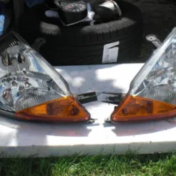 Dwie używane lampy samochodowe, lewa i prawa, z pomarańczowymi kierunkowskazami, leżą na białej powierzchni, w tle opona i przyrząd pomiarowy.