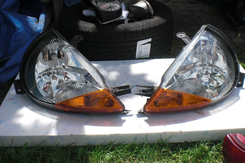 Dwie używane lampy samochodowe, lewa i prawa, z pomarańczowymi kierunkowskazami, leżą na białej powierzchni, w tle opona i przyrząd pomiarowy.