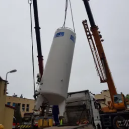 Dwa dźwigi podnoszą pionowy, biały zbiornik Linde z platformy ciężarówki, widoczny budynek w tle.