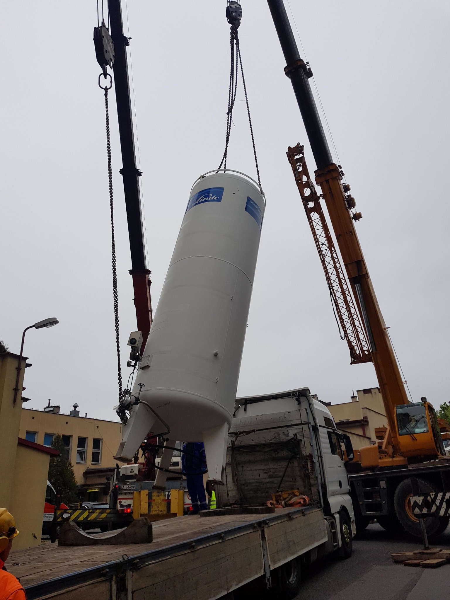 Dwa dźwigi podnoszą pionowy, biały zbiornik Linde z platformy ciężarówki, widoczny budynek w tle.