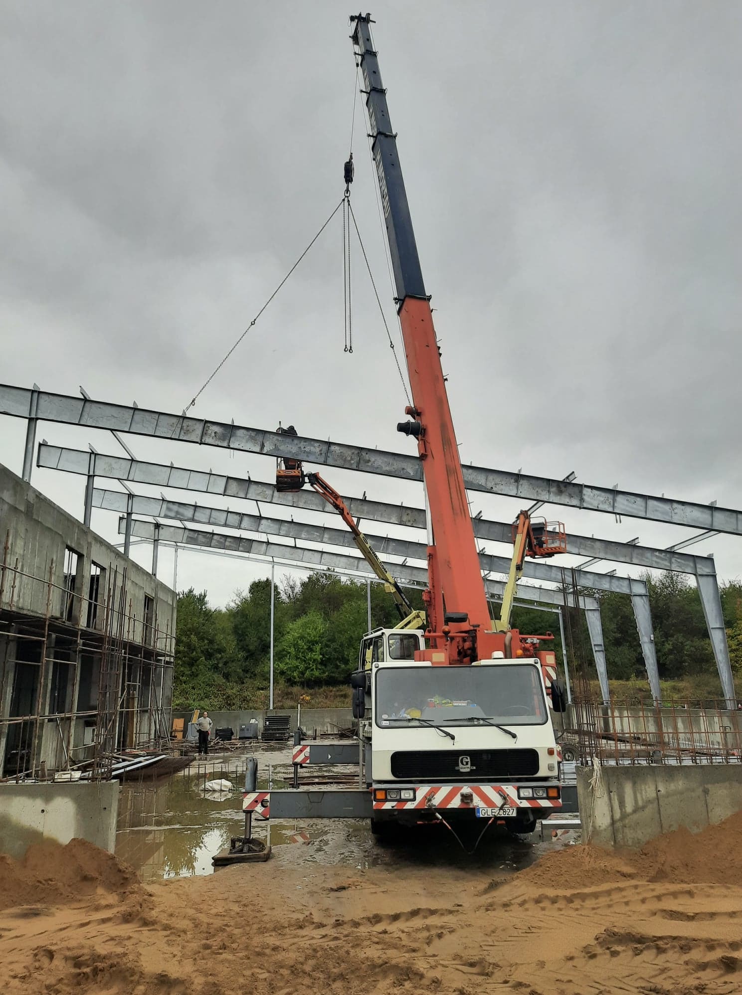 Mobilny żuraw podnosi stalową belkę konstrukcyjną na budowie, widoczne podnośniki koszowe, teren podmokły po deszczu, pochmurne niebo w tle.