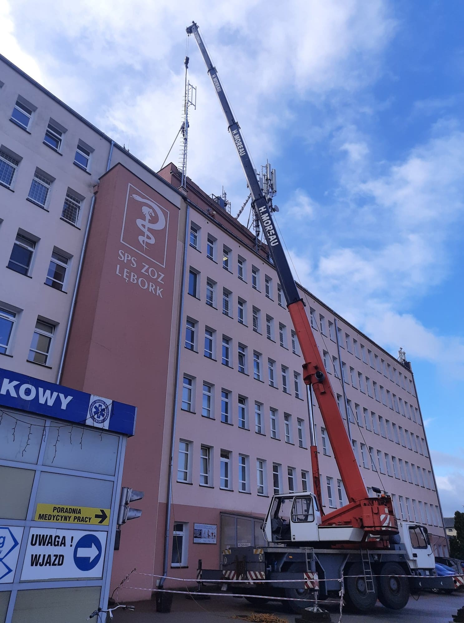 Czerwono-biały żuraw samochodowy H. Moreau unosi antenę telekomunikacyjną na dach budynku SPS ZOZ Lębork, widoczne błękitne niebo z chmurami.