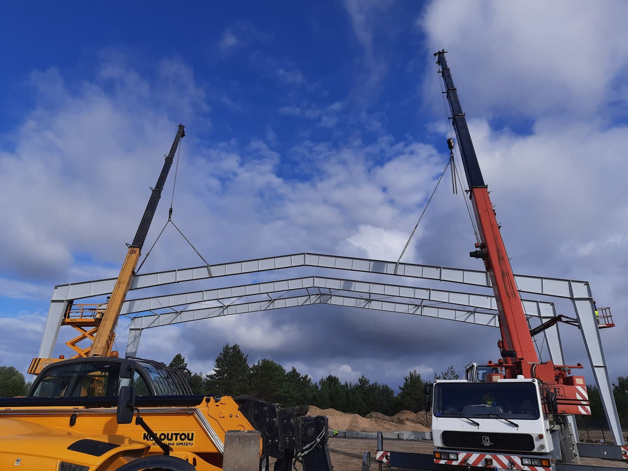 Montaż konstrukcji stalowej hali za pomocą dwóch dźwigów z wysięgnikami teleskopowymi na tle błękitnego nieba z chmurami.