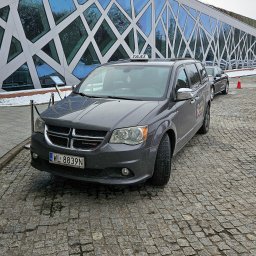 TAXI SEROCK MITER - Taksówka 24h - Postój Taxi - Szary minivan Dodge z napisem TAXI na dachu, zaparkowany na brukowanej nawierzchni przed budynkiem o nowoczesnej architekturze z geometrycznymi wzorami na szklanej fasadzie.
