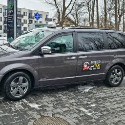 TAXI SEROCK MITER - Taksówka 24h - Postój Taxi - Szary minivan z oznaczeniami taksówki, zaparkowany na chodniku z kostki brukowej z resztkami śniegu, widoczny znak firmy taksówkarskiej 'MITER TAXI' na boku pojazdu.