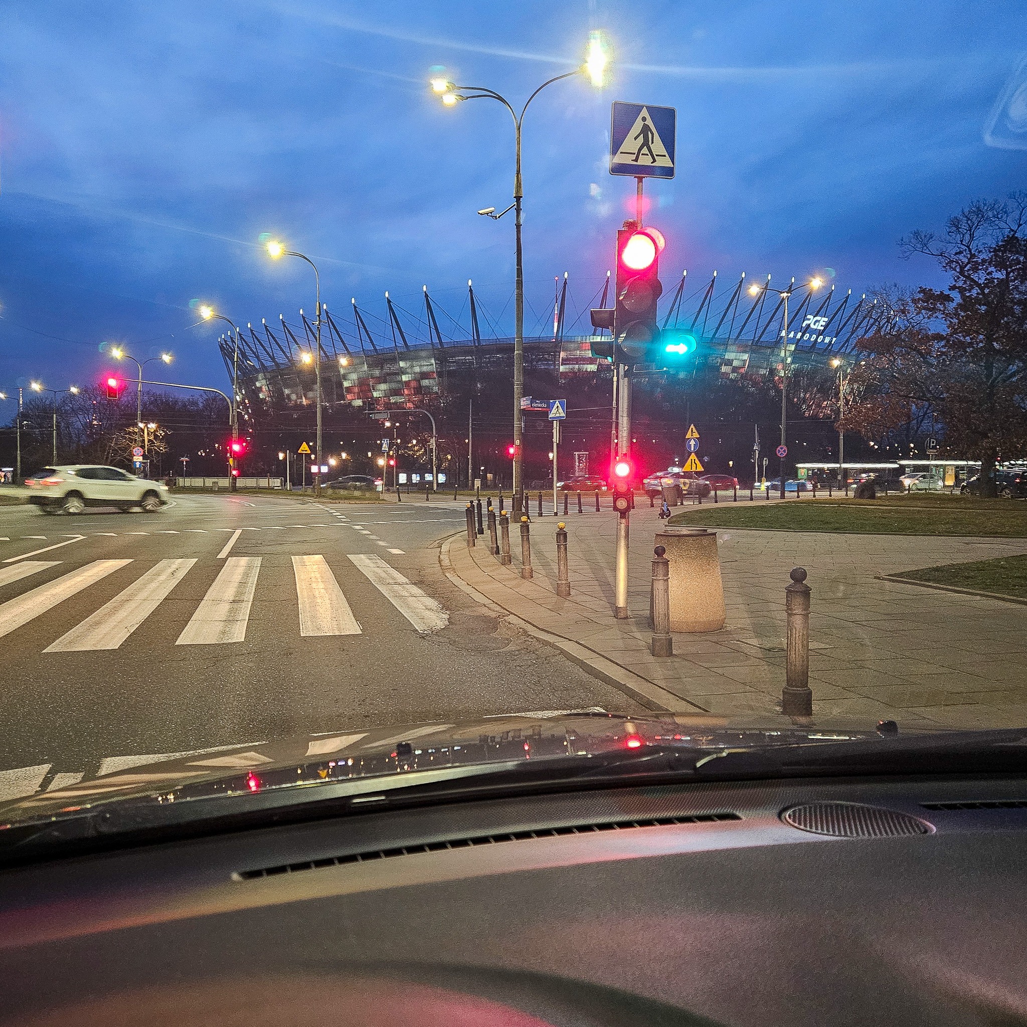 Widok z wnętrza samochodu na skrzyżowanie z przejściem dla pieszych, sygnalizacją świetlną i Stadionem Narodowym w tle, wieczorową porą.