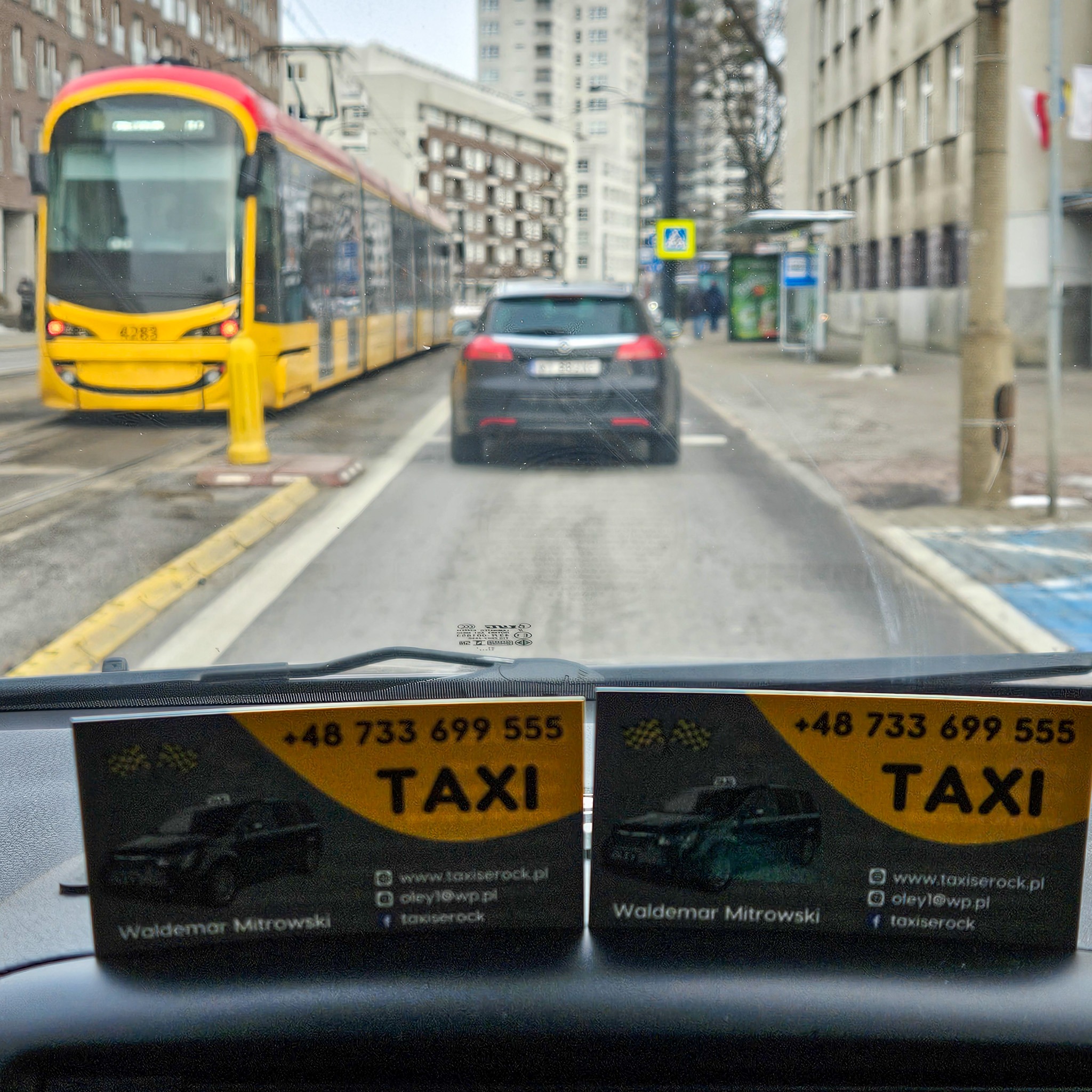 Widok z wnętrza taksówki na ulicę miejską, z tramwajem i samochodem w tle, wizytówki taksówkarza na desce rozdzielczej z numerem telefonu i nazwą firmy.