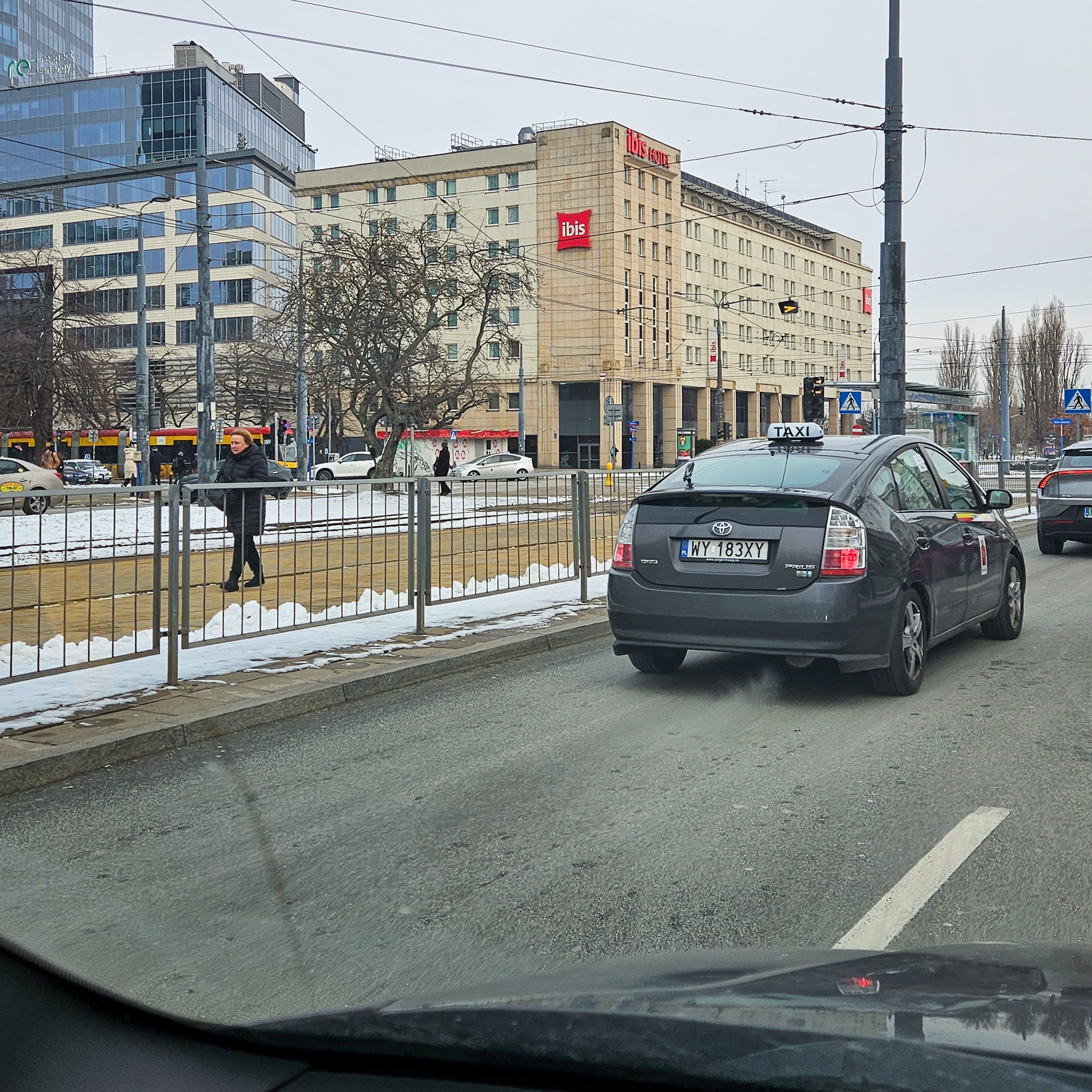 Szary samochód Toyota Prius z napisem TAXI na dachu, stojący na ulicy w Serocku, z widocznym budynkiem Ibis Hotel w tle.