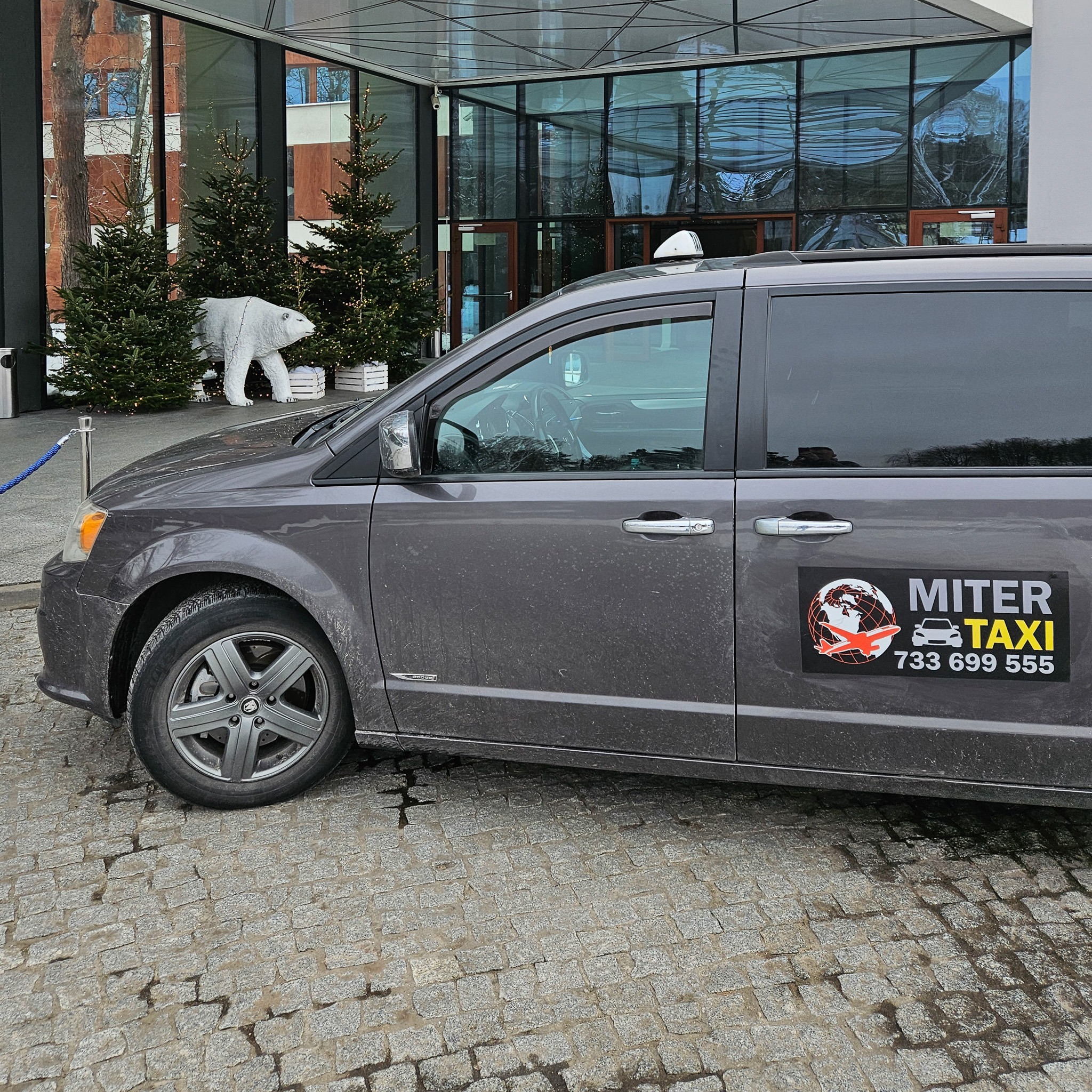 Szary minivan taxi z naklejką firmy 'MITER TAXI' zaparkowany na brukowanej nawierzchni przed nowoczesnym budynkiem z choinkami i dekoracyjnym białym niedźwiedziem.