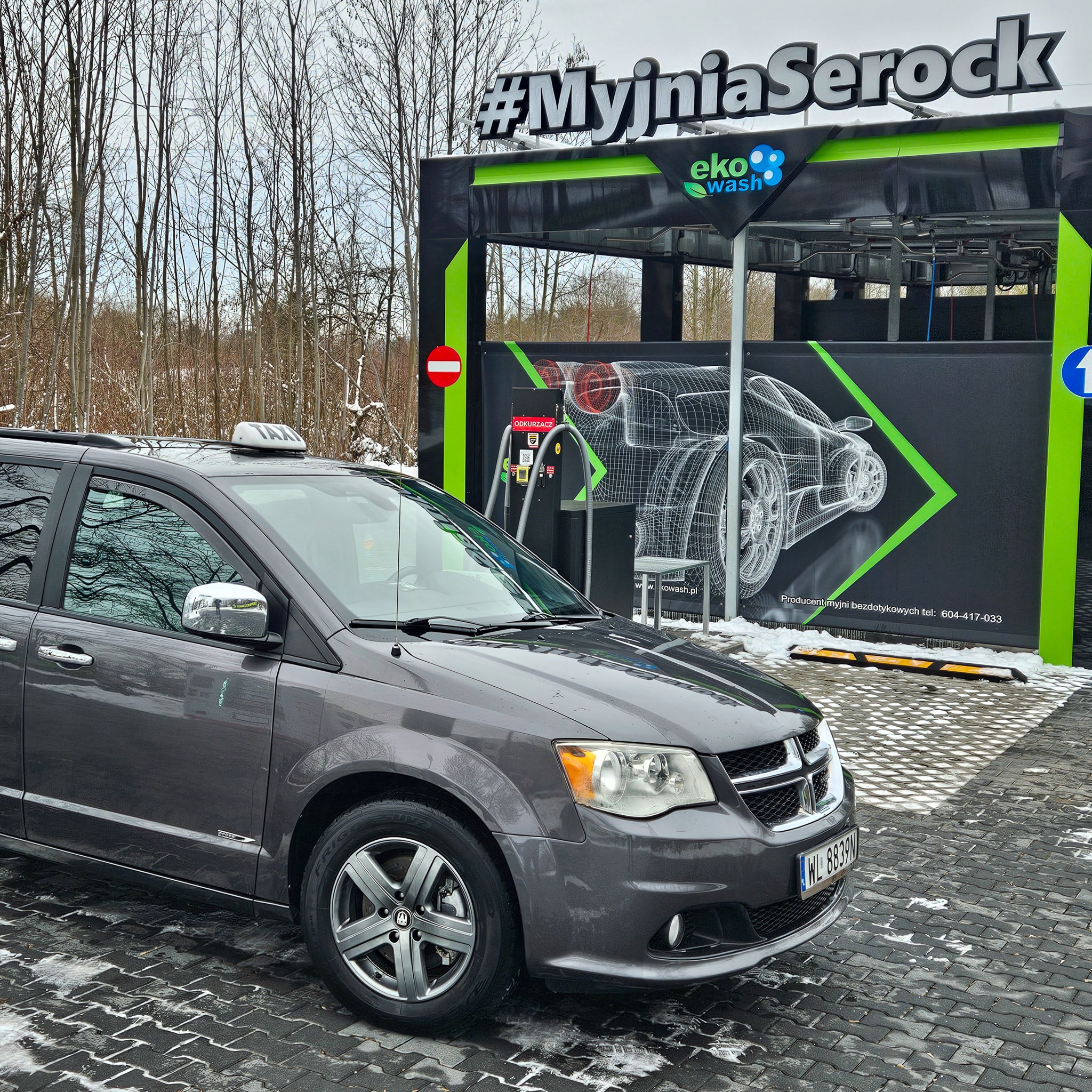 Szary minivan z taksometrem na dachu przed nowoczesną, bezdotykową myjnią samochodową Eko Wash z napisem #MyjniaSerock, widoczne drzewa w tle.