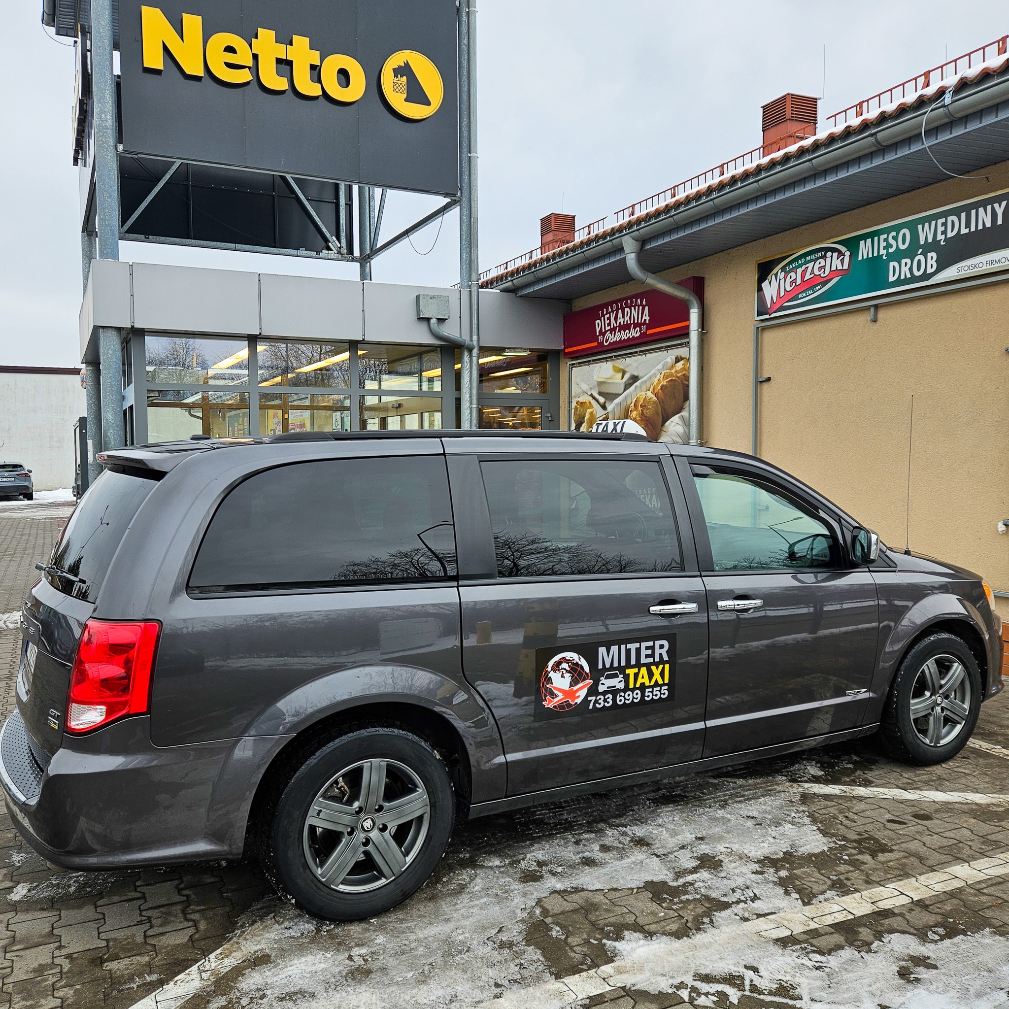 Szary minivan z naklejką 'Miter Taxi' zaparkowany przed sklepem Netto i piekarnią Oskroba w zimowy dzień.