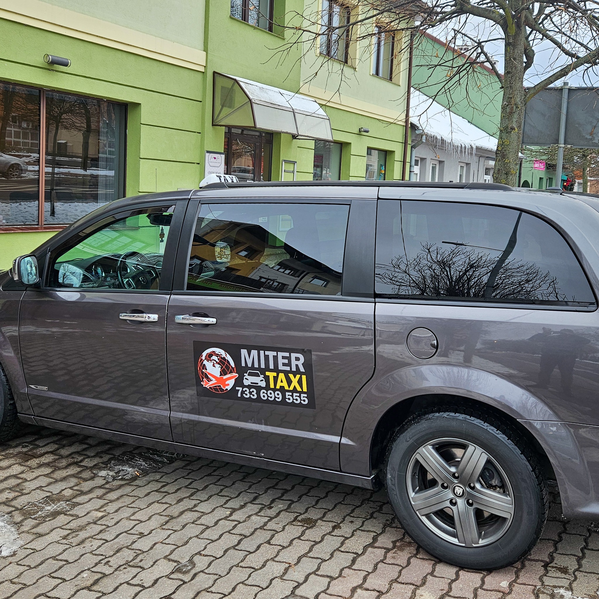 Szary minivan z naklejką reklamową taksówki Miter Taxi z numerem telefonu 733 699 555, zaparkowany na chodniku z kostki brukowej przed zielonym budynkiem w pochmurny dzień.