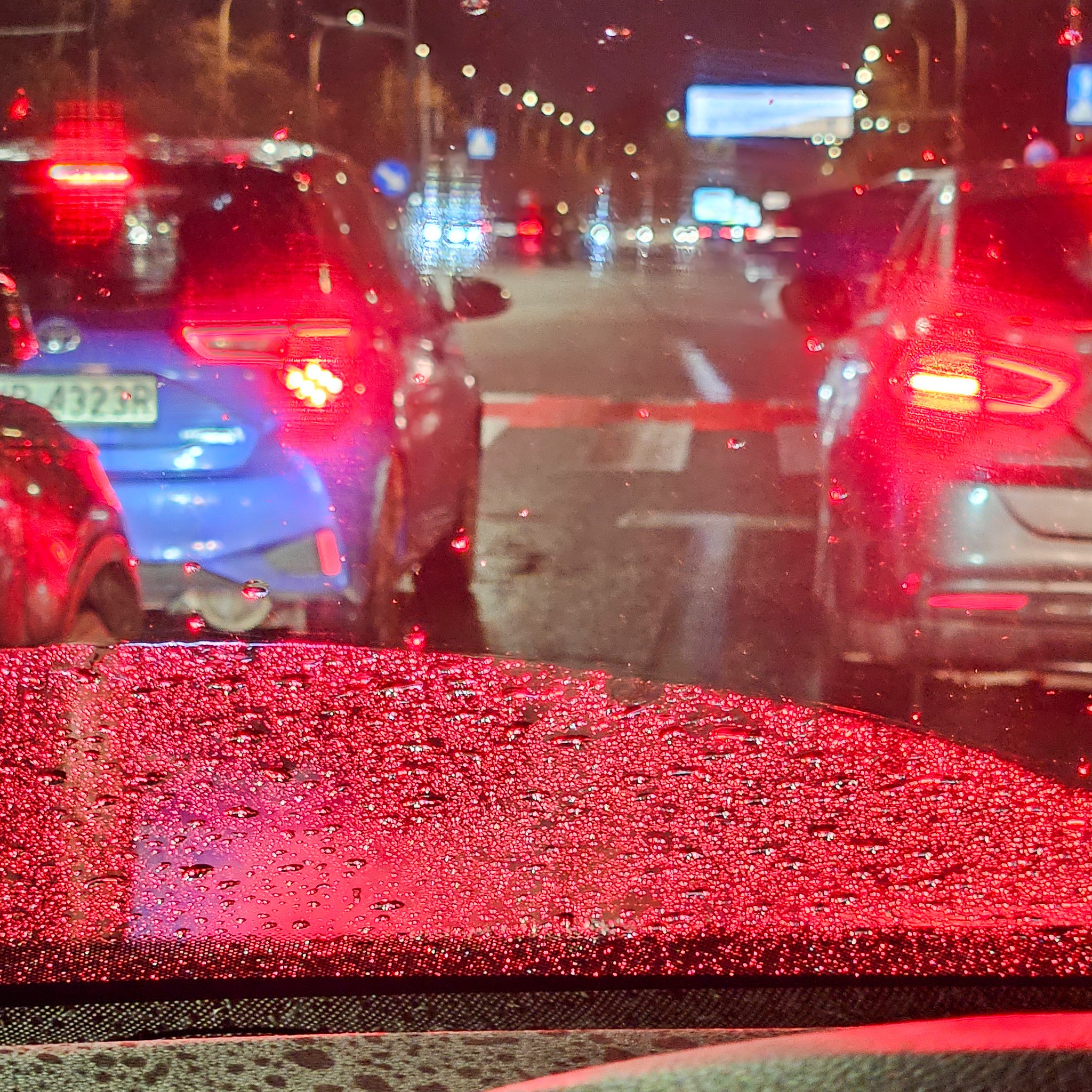 Widok z wnętrza samochodu na ulicę w nocy, przez zaparowaną szybę z kroplami deszczu, w tle światła samochodów i budynków.