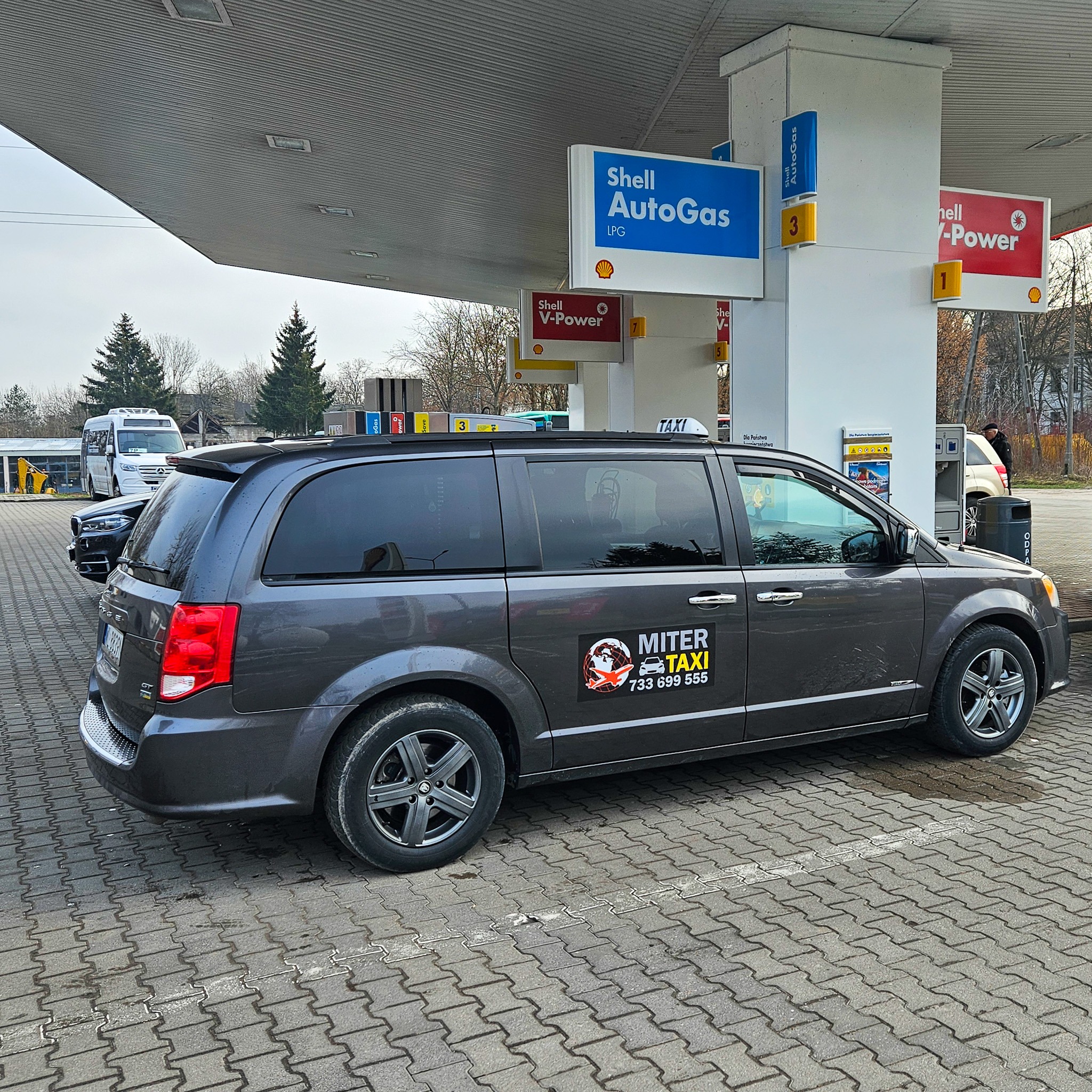 Szary minivan z naklejką firmy taksówkarskiej Miter Taxi parkuje na stacji benzynowej Shell, widoczny dystrybutor LPG i V-Power.