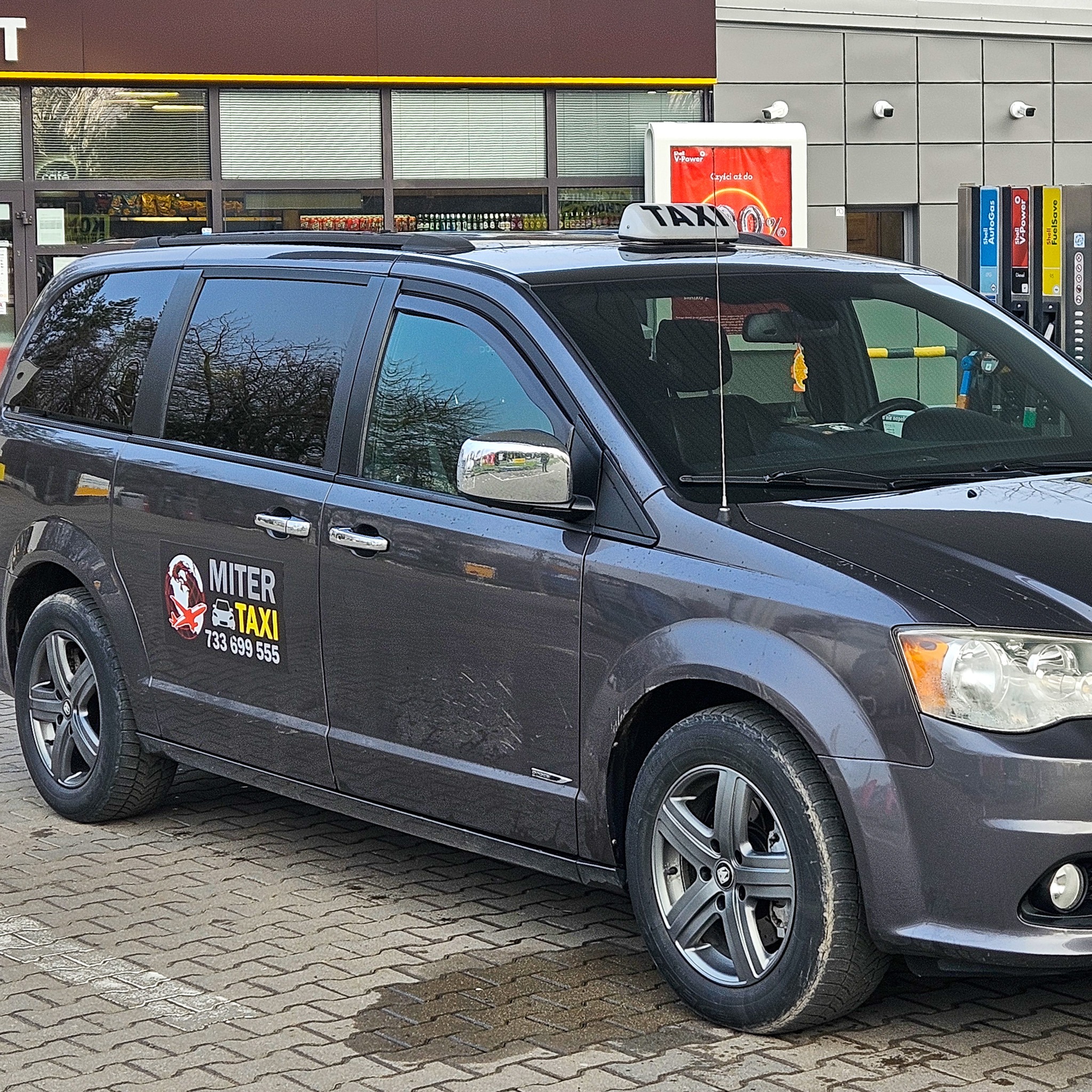 Szary minivan taxi z logo 'MITER TAXI' na tle stacji benzynowej Shell, z widocznym szyldem 'TAXI' na dachu