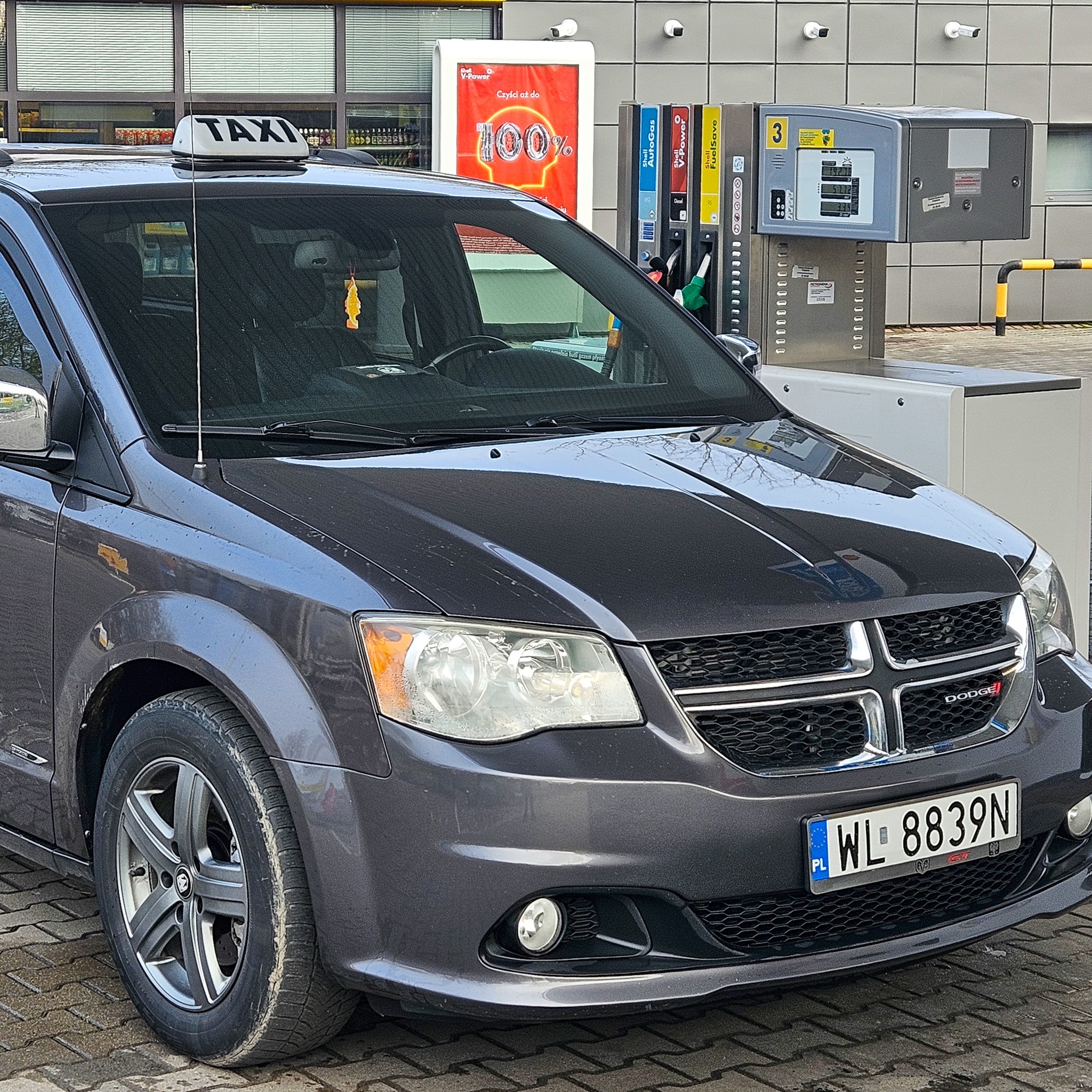 Szary minivan z napisem TAXI na dachu stojący przy dystrybutorze paliwa na stacji benzynowej z widoczną polską tablicą rejestracyjną.