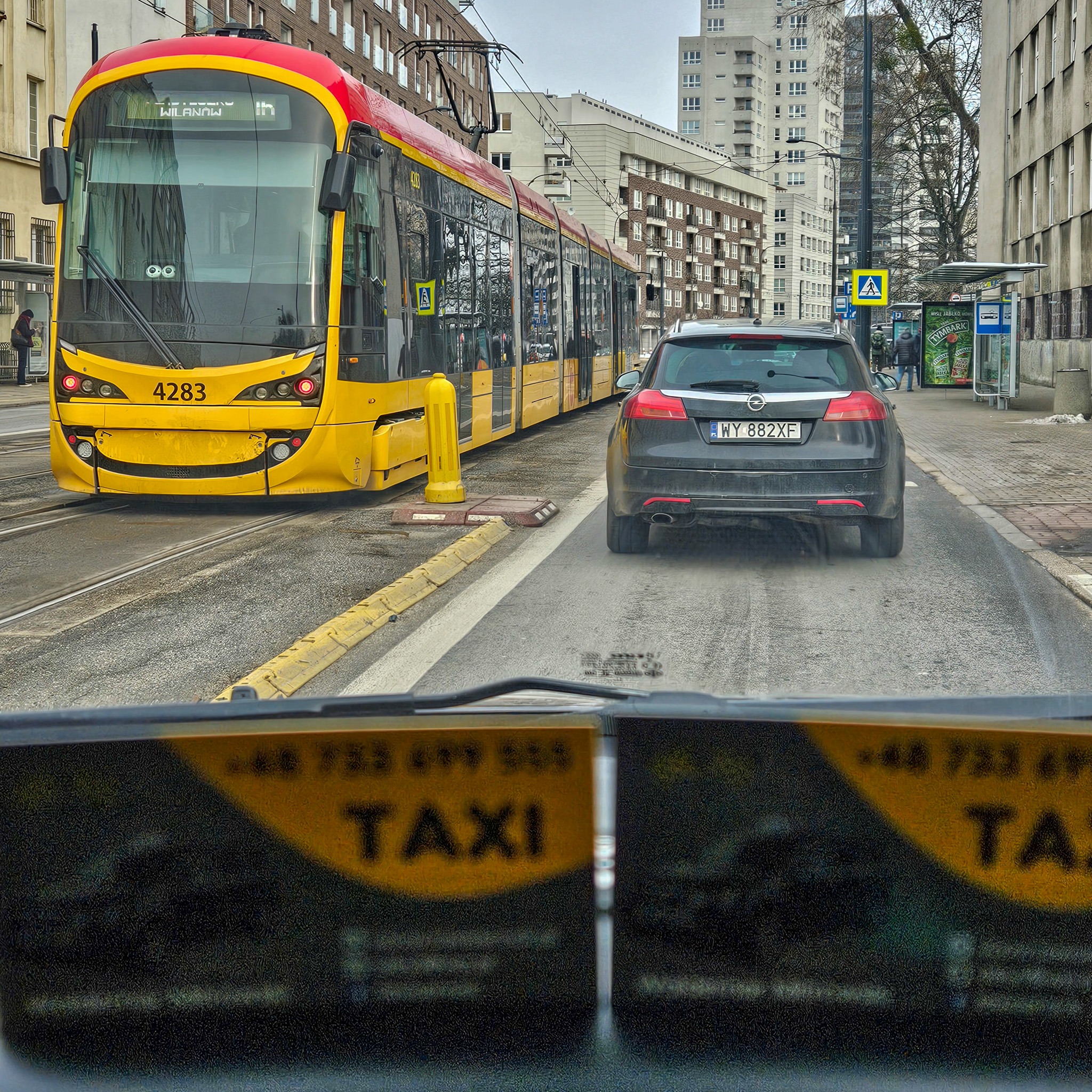 Widok z wnętrza taksówki na ulicę miejską z tramwajem i samochodem osobowym, w tle budynki.