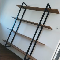 JAK-MET Jakubowski Dawid - Ręcznie robiona, minimalistyczna półka ścienna z drewna i czarnej stali, z czterema poziomami, zamontowana na białej ścianie, widoczny parkiet na podłodze i gniazdko elektryczne