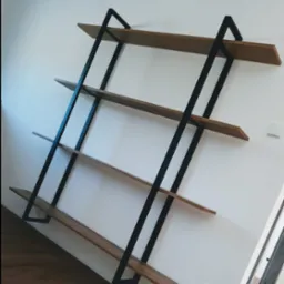 Ręcznie robiona, minimalistyczna p&oacute;łka ścienna z drewna i czarnej stali, z czterema poziomami, zamontowana na białej ścianie, widoczny parkiet na podłodze i gniazdko elektryczne