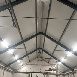 JAK-MET Jakubowski Dawid - Wnętrze hali w trakcie budowy lub remontu z widoczną konstrukcją stalową dachu, rusztowaniami, ścianami z białego bloczka i pracownikiem przy maszynie.