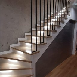 JAK-MET Jakubowski Dawid - Nowoczesne schody drewniane z podświetleniem LED i minimalistyczną, czarną balustradą z profili kwadratowych, na tle ściany z efektem betonu.