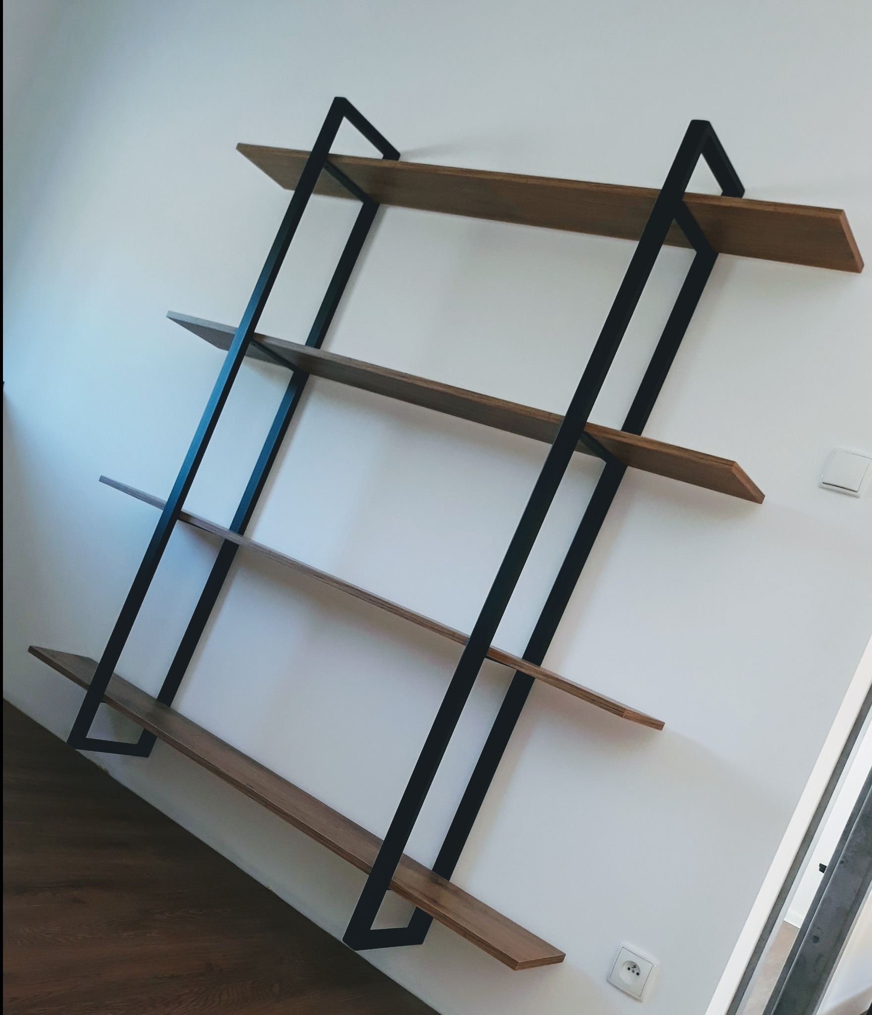 Ręcznie robiona, minimalistyczna półka ścienna z drewna i czarnej stali, z czterema poziomami, zamontowana na białej ścianie, widoczny parkiet na podłodze i gniazdko elektryczne