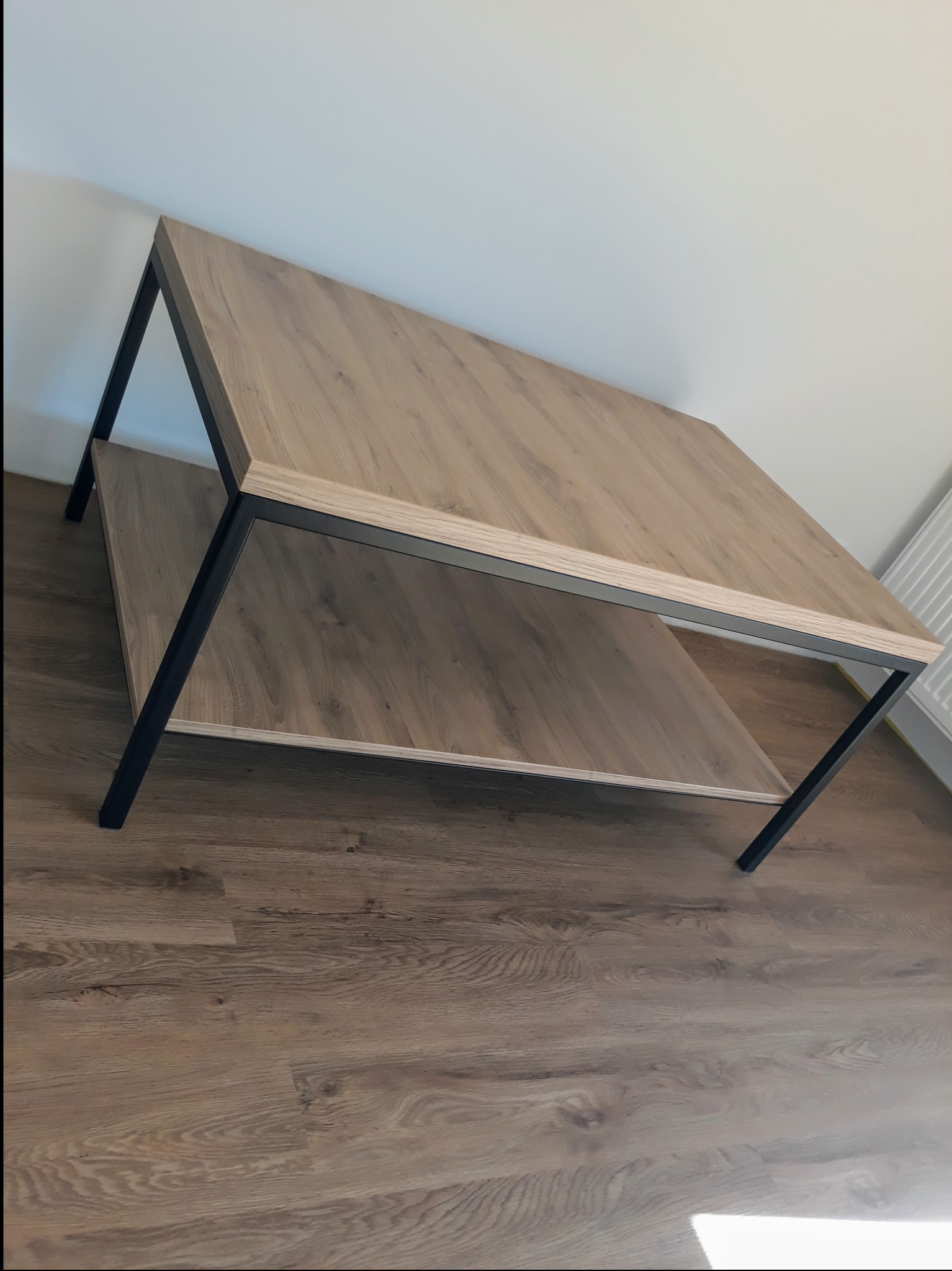 Stolik kawowy z drewnianym blatem i czarną metalową ramą, widoczna dolna półka, ustawiony na drewnianej podłodze, kątowe ujęcie.