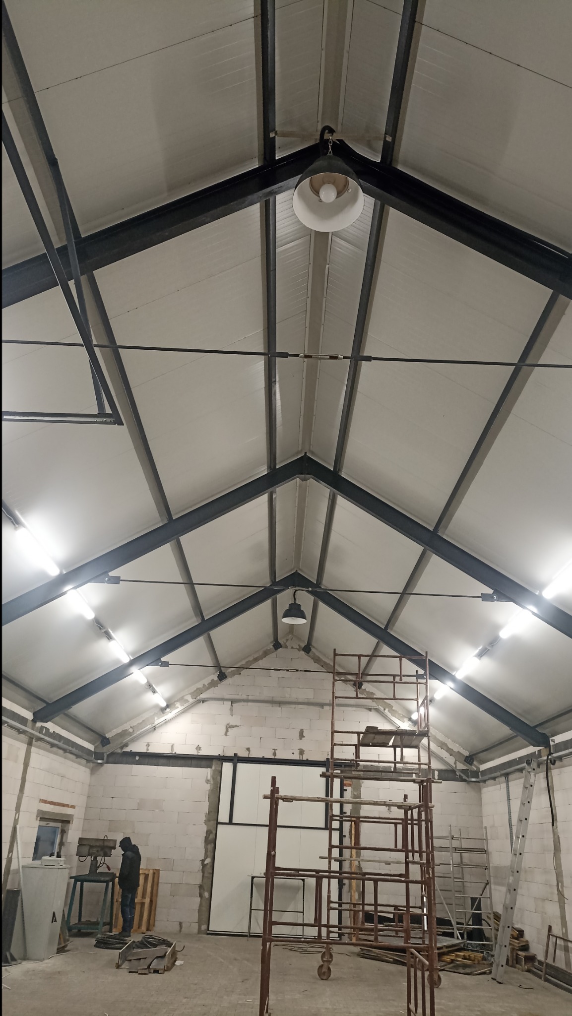 Wnętrze hali produkcyjnej w trakcie budowy z widoczną konstrukcją stalową dachu, oświetleniem, rusztowaniami i osobą przy maszynie do obróbki metalu.