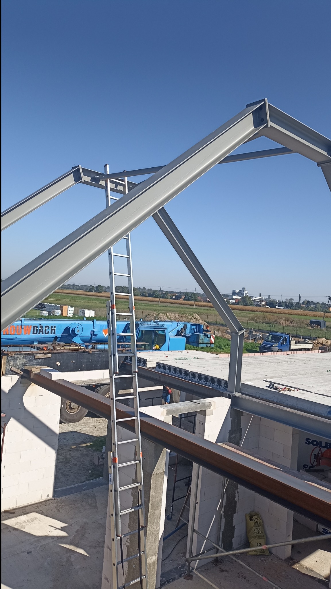 Montaż stalowej konstrukcji dachu budynku w trakcie budowy, widoczny dźwig budowlany w tle, drabina aluminiowa oparta o ścianę z betonu komórkowego.