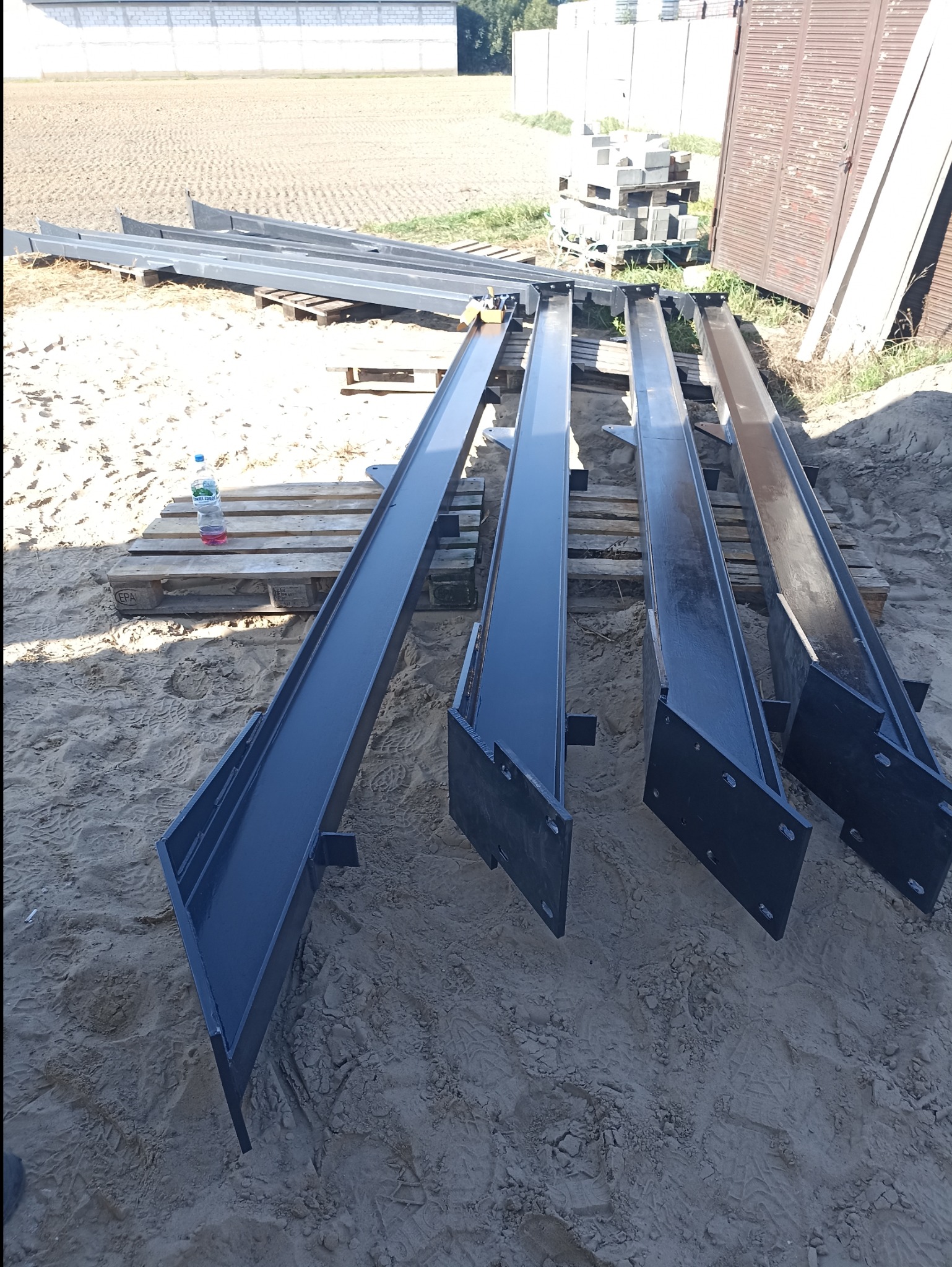 Czarne, pomalowane profile stalowe o przekroju ceownika, leżące na drewnianych paletach na piaszczystym podłożu. Widoczna butelka wody mineralnej. W tle budynek z białej cegły i elementy...