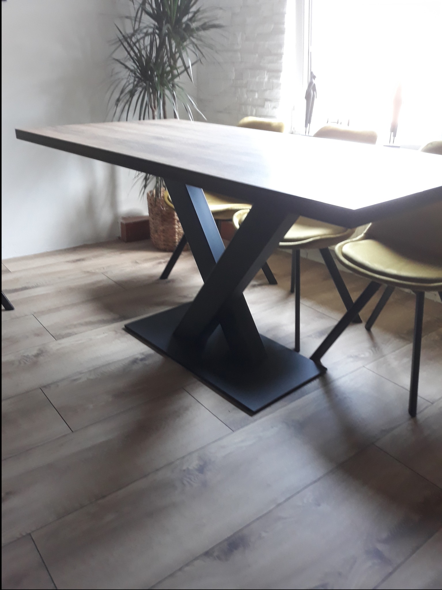 Stół z drewnianym blatem i czarną, metalową podstawą w kształcie litery X, ustawiony na drewnianej podłodze w jasnym wnętrzu.