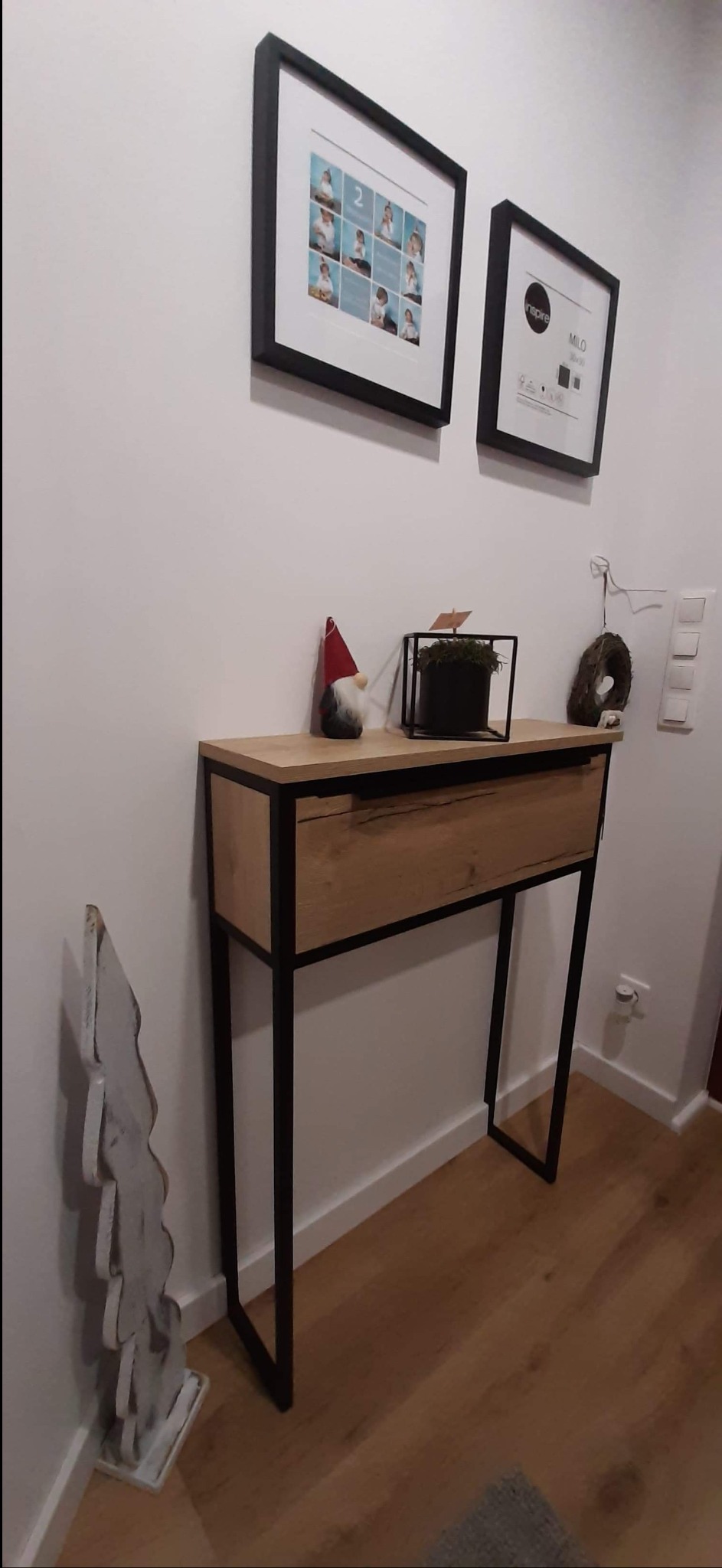 Konsola z szufladą, drewniany blat i czarne metalowe nogi, dekoracje na blacie, zdjęcia w czarnych ramkach na ścianie.