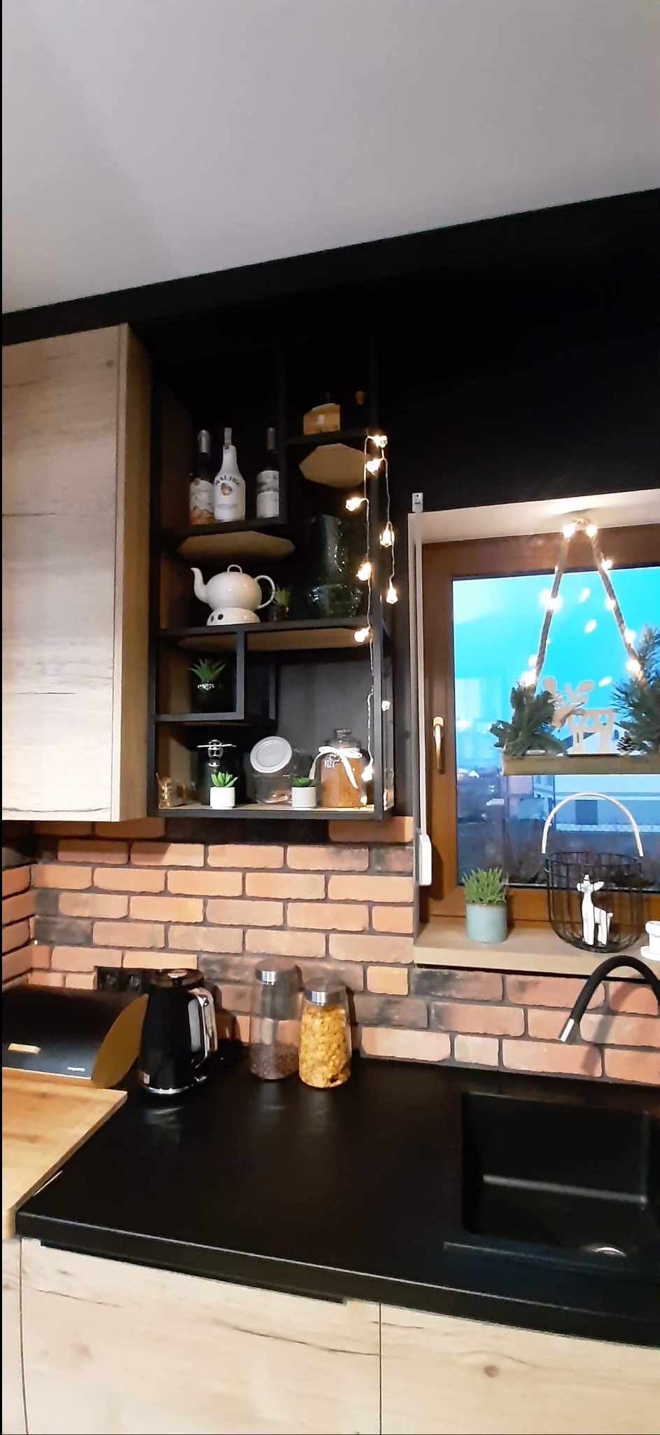 Kuchenna zabudowa z otwartymi półkami z czarnej stali, ceglana ściana nad blatem, dekoracje okienne z motywem świątecznym, czarny zlew i bateria.