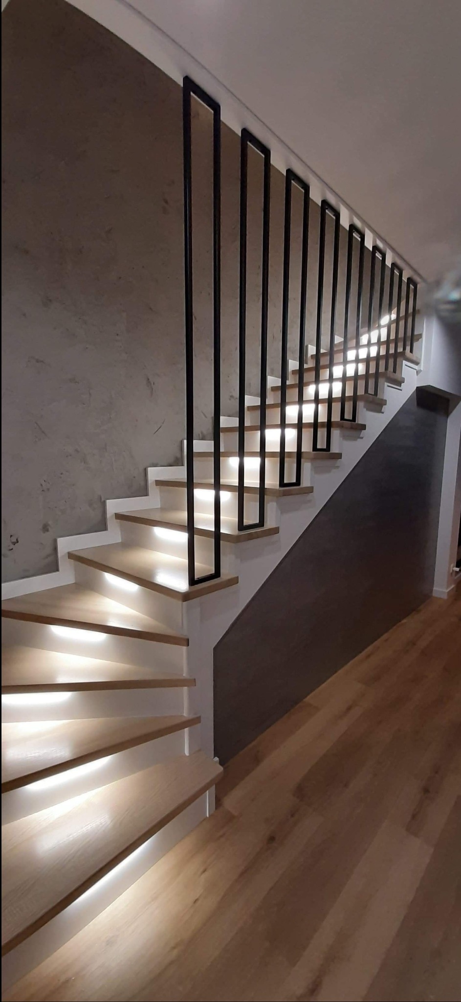 Nowoczesne schody drewniane z podświetleniem LED i minimalistyczną, czarną balustradą z profili kwadratowych, na tle ściany z efektem betonu.