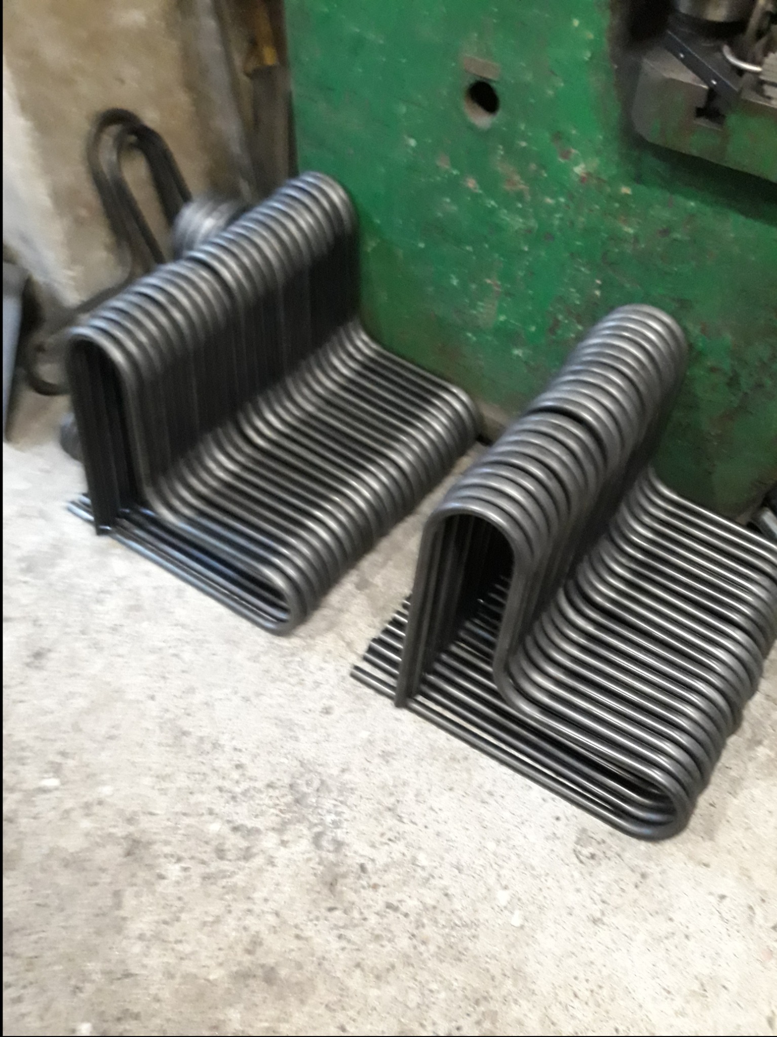 Stosy giętych, metalowych rurek w kształcie litery U, ułożone obok zielonej maszyny warsztatowej.