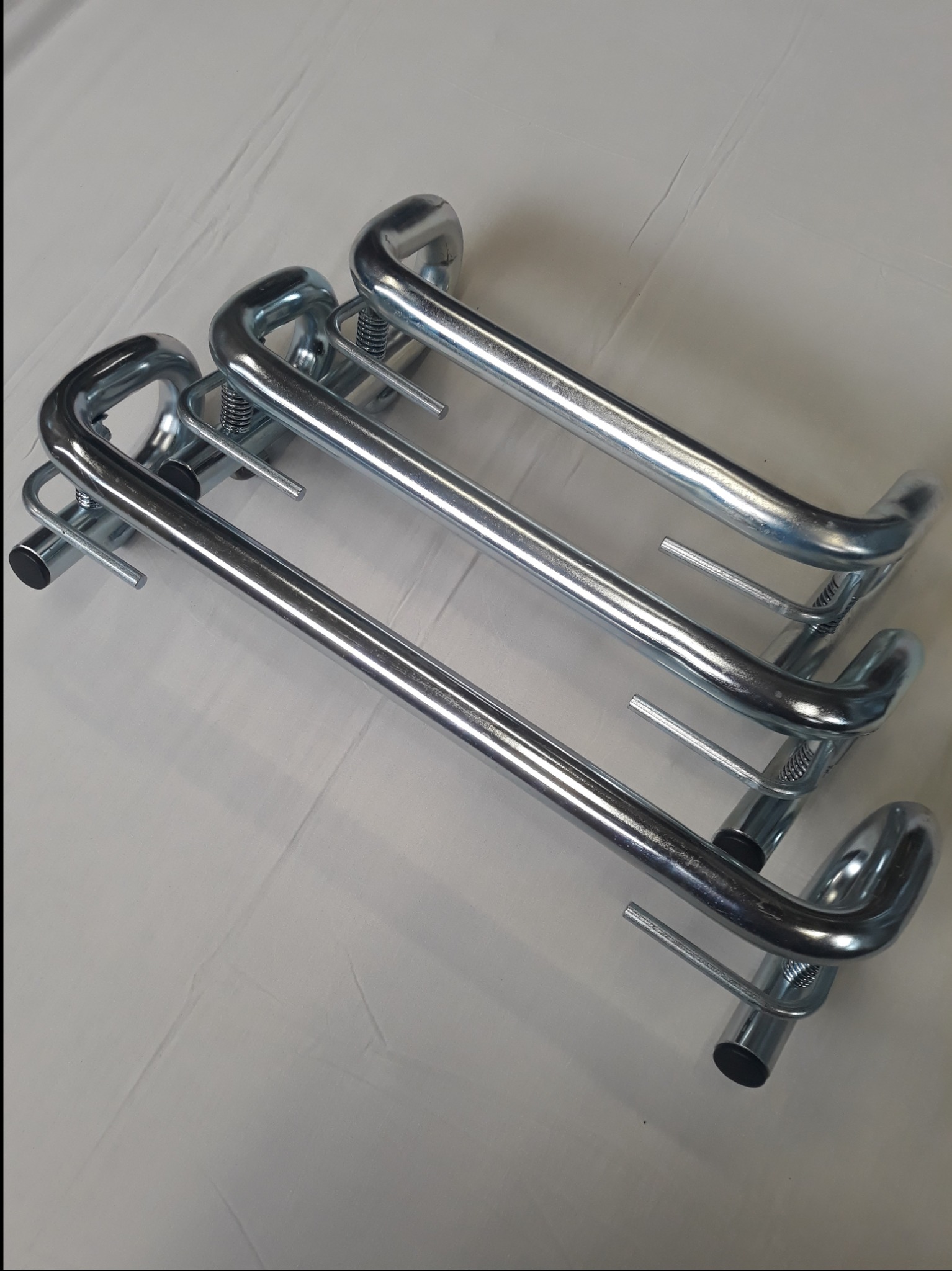 Trzy chromowane, gięte rury z elementami sprężynowymi, ułożone równolegle na jasnym tle, widoczne ślady obróbki metalu.
