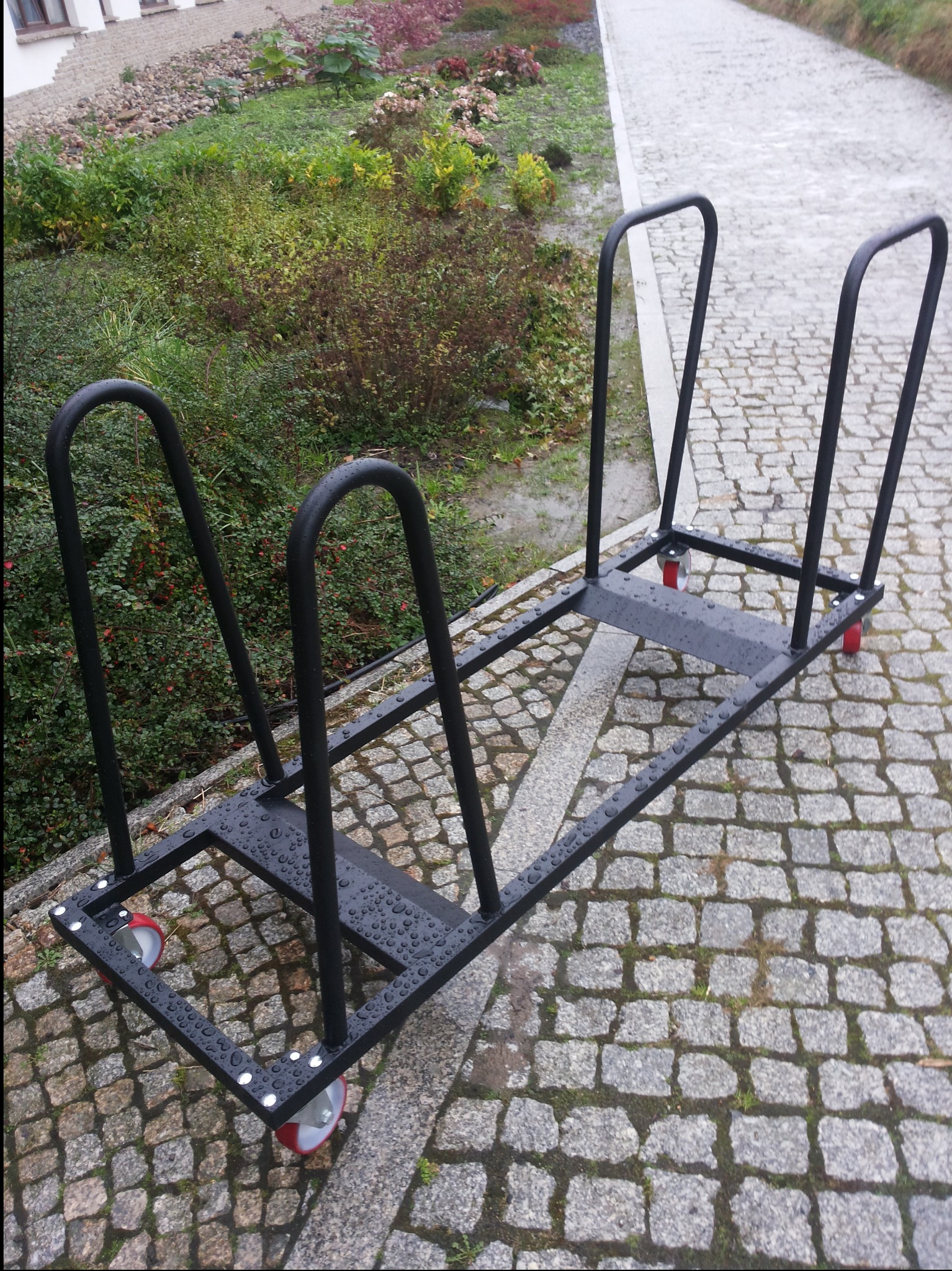 Metalowy wózek transportowy z czarnych profili z czterema czerwonymi kółkami, stojący na kostce brukowej, pokryty kroplami deszczu.