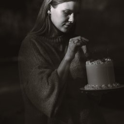 Anna Rucińska - Monochromatyczne ujęcie kobiety w swetrze, dekorującej tort z białym kremem i wisienkami na talerzu.