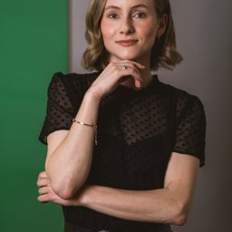 Anna Rucińska - Portret kobiety w czarnej bluzce w kropki, z ręką pod brodą, na tle zielono-szarym. Widoczny pierścionek i złota bransoletka.