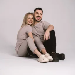 Uśmiechnięta blondynka i brunet siedzą na podłodze w dopasowanych beżowych uniformach medycznych i obuwiu typu Crocs, pozują na jasnym tle.