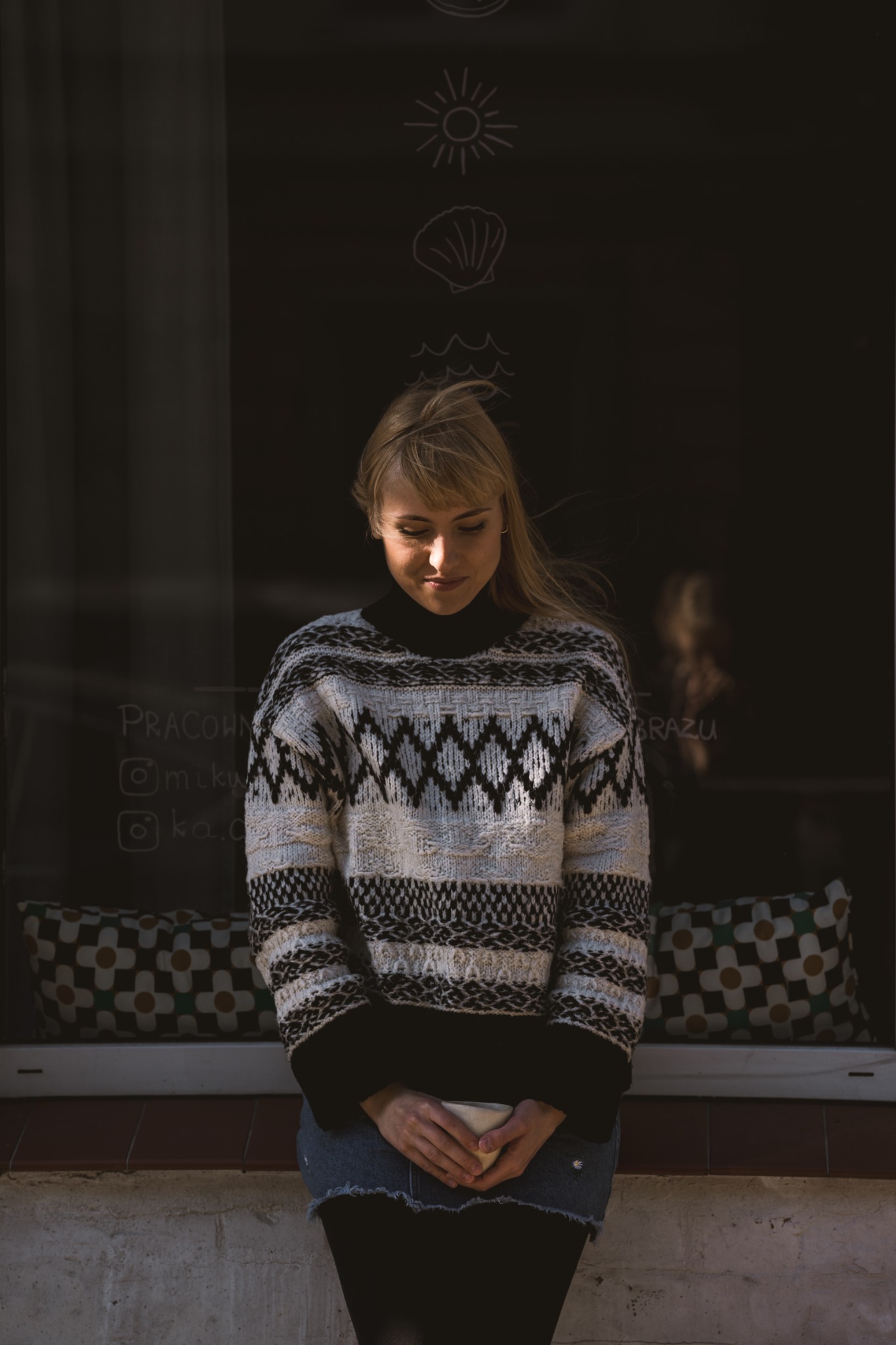 Kobieta w swetrze w geometryczne wzory, stojąca przed oknem z rysunkami kredą, trzymająca kubek w dłoniach, z delikatnym uśmiechem.