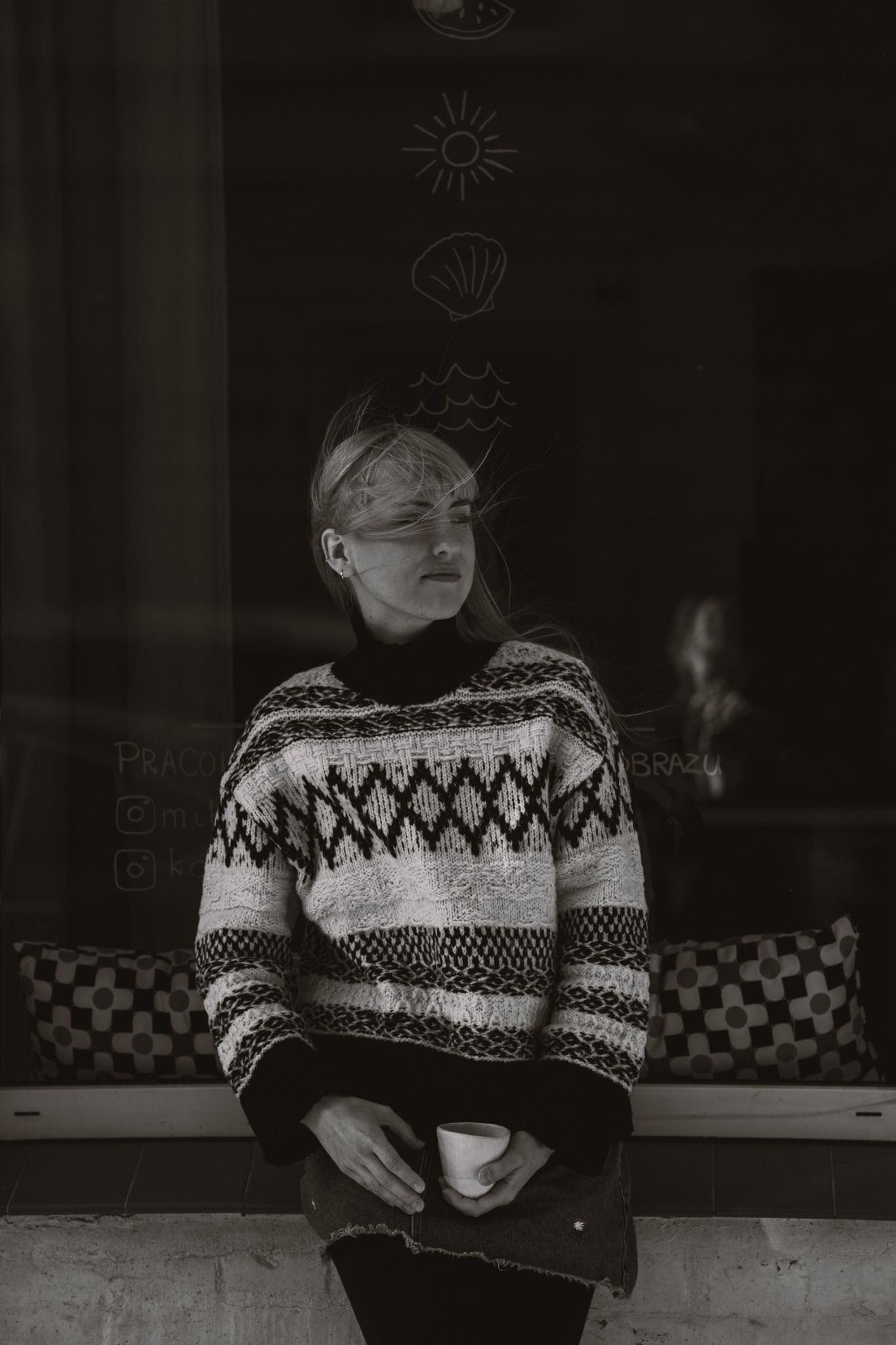 Portret kobiety w swetrze z wzorem, trzymającej filiżankę na tle szyby z rysunkami kredą, w czarno-białej tonacji.