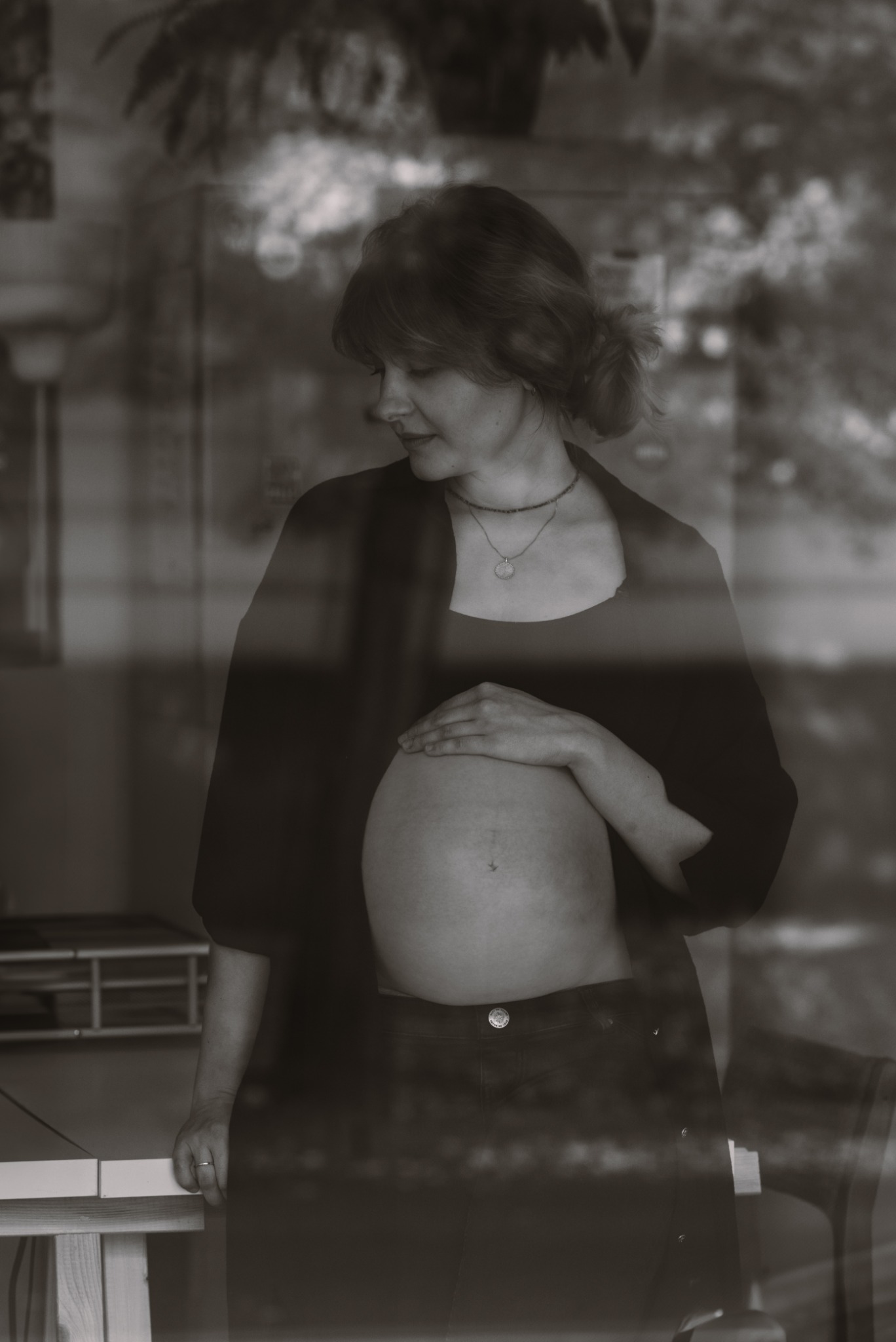 Czarno-białe portretowe ujęcie kobiety w ciąży, delikatnie opierającej dłoń na brzuchu, ubrana w ciemną koszulę i spodnie, widoczna przez szybę z odbiciami otoczenia.