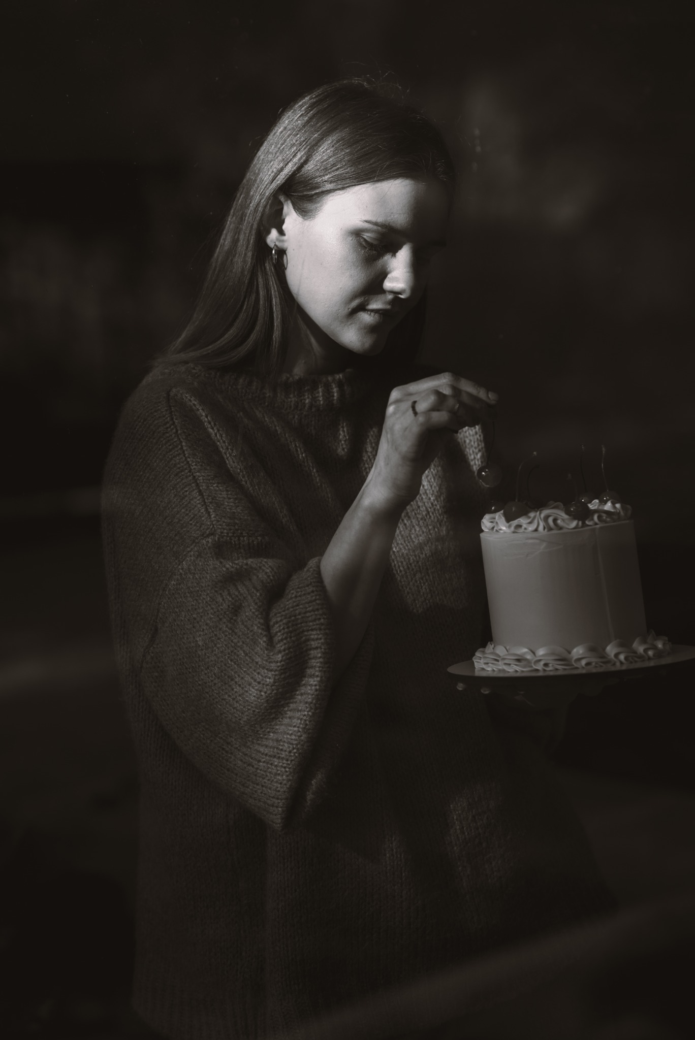 Monochromatyczne ujęcie kobiety w swetrze, dekorującej tort z białym kremem i wisienkami na talerzu.