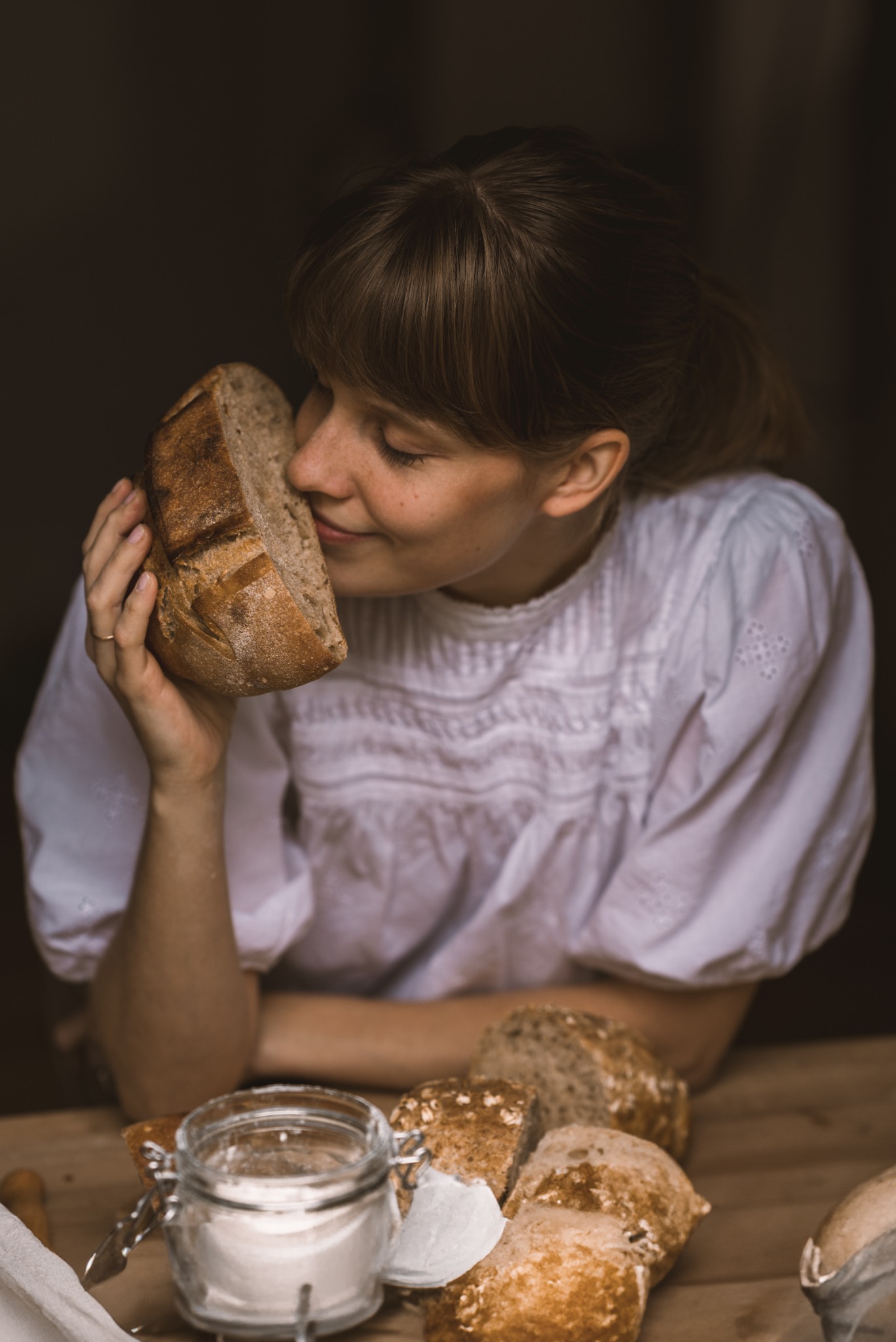 Kobieta z piegami w białej bluzce wącha kromkę chleba na drewnianym stole, obok słoik z mąką i kilka bułek.