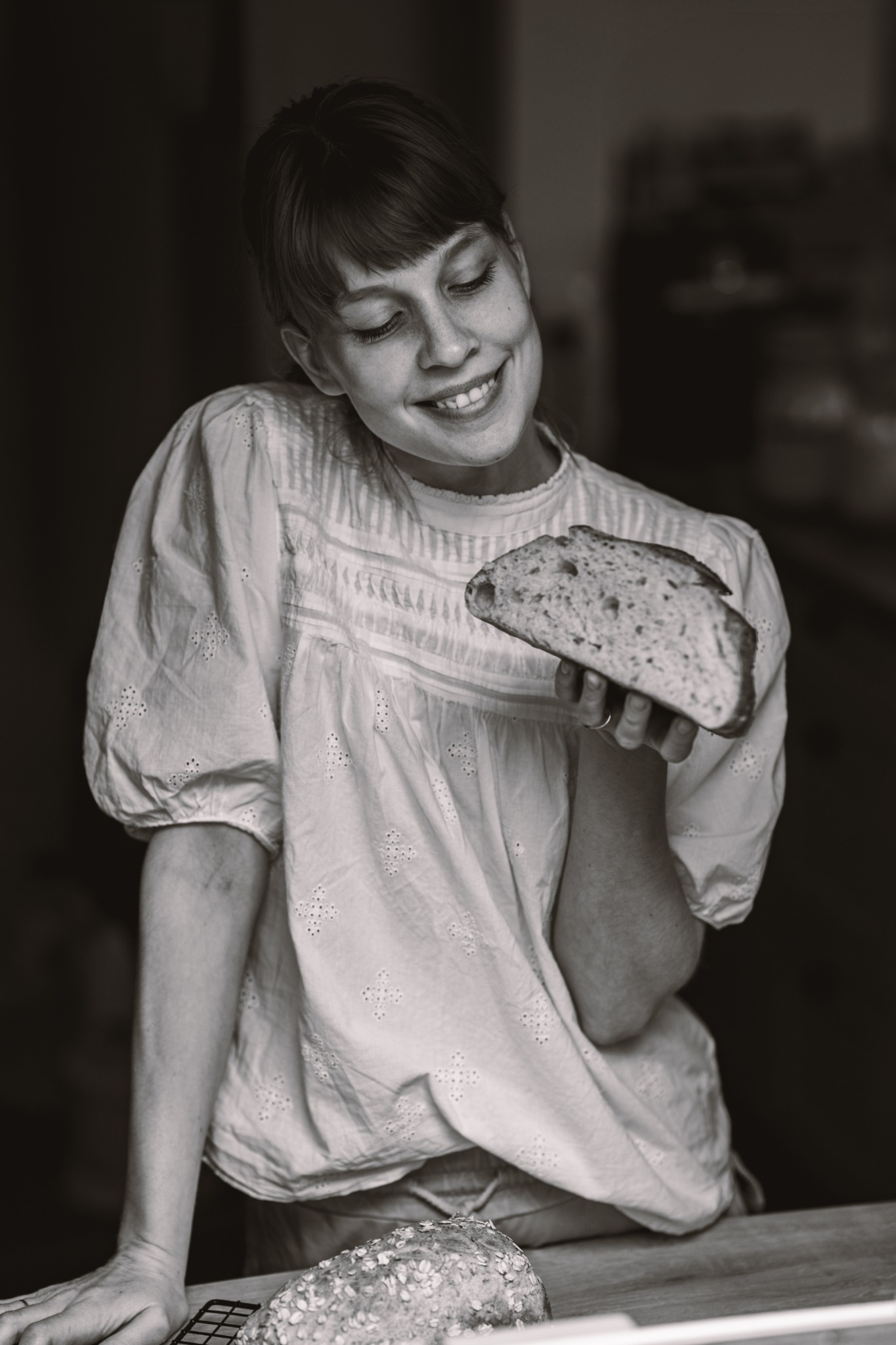 Uśmiechnięta kobieta w bluzce z haftem trzyma kromkę chleba na tle ciemnego wnętrza; na stole leży bochenek chleba na metalowej kratce.