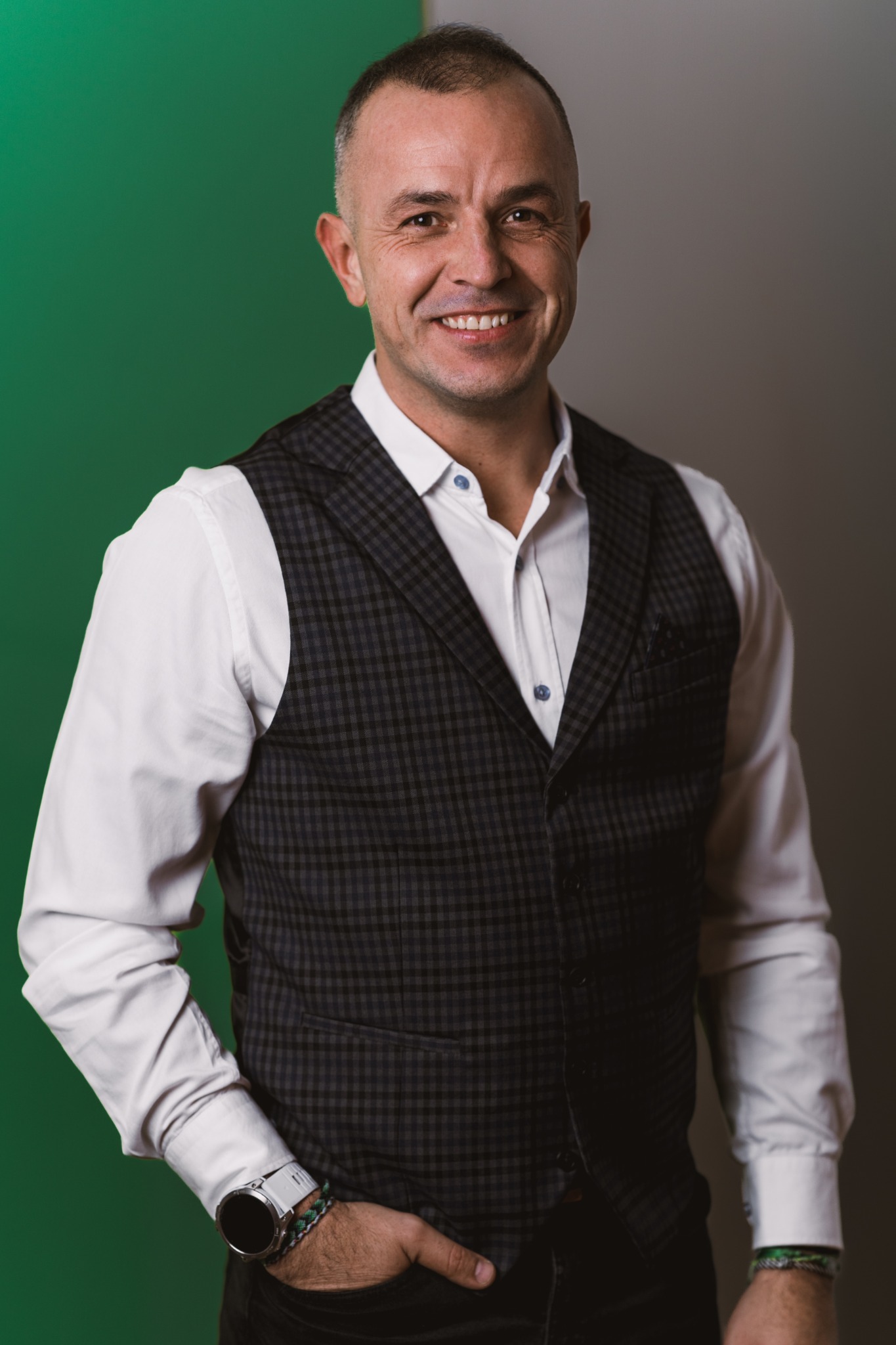 Uśmiechnięty mężczyzna w szarej kraciastej kamizelce i białej koszuli, z zegarkiem na ręku, na tle zielono-szarego tła.