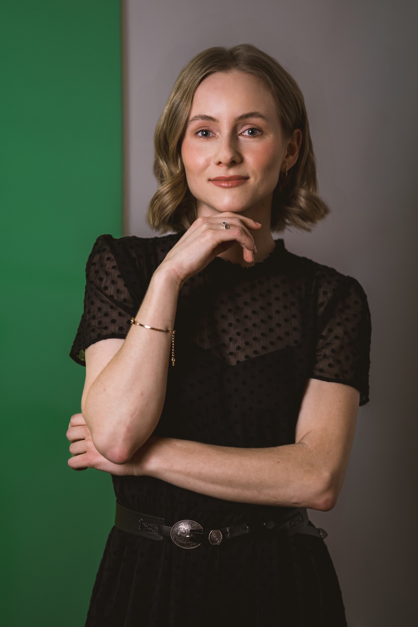Portret kobiety w czarnej bluzce w kropki, z ręką pod brodą, na tle zielono-szarym. Widoczny pierścionek i złota bransoletka.