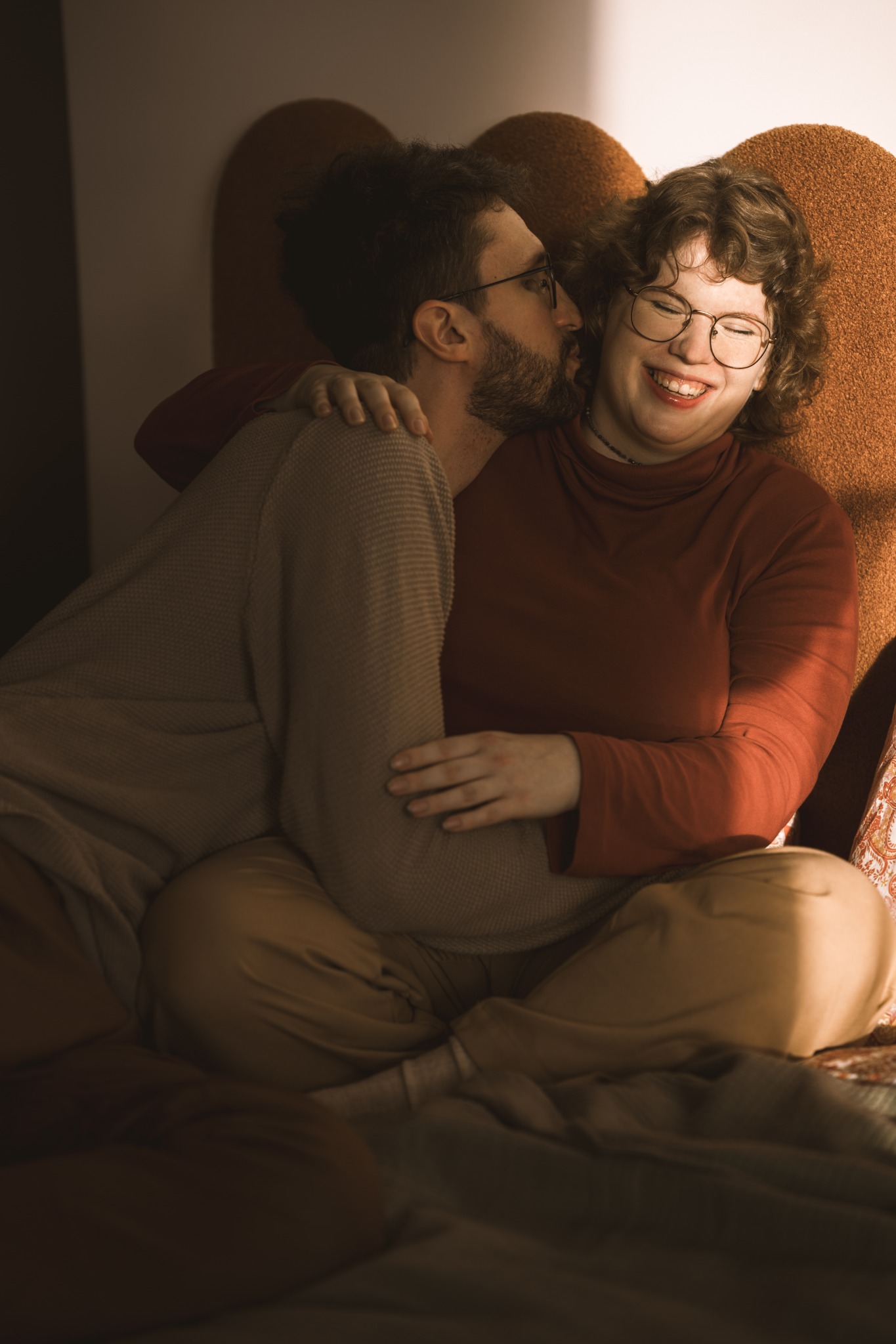 Uśmiechnięta kobieta w okularach, siedząca na łóżku, obejmowana przez mężczyznę, który ją całuje w policzek, ciepłe światło w tle.