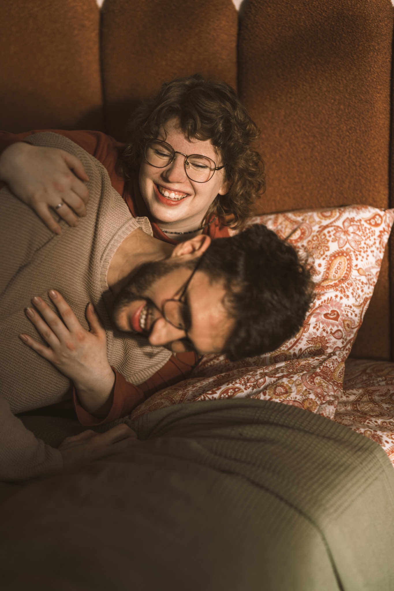 Uśmiechnięta para w okularach, obejmująca się na łóżku z wzorzystą pościelą, ciepłe oświetlenie, brązowe tło